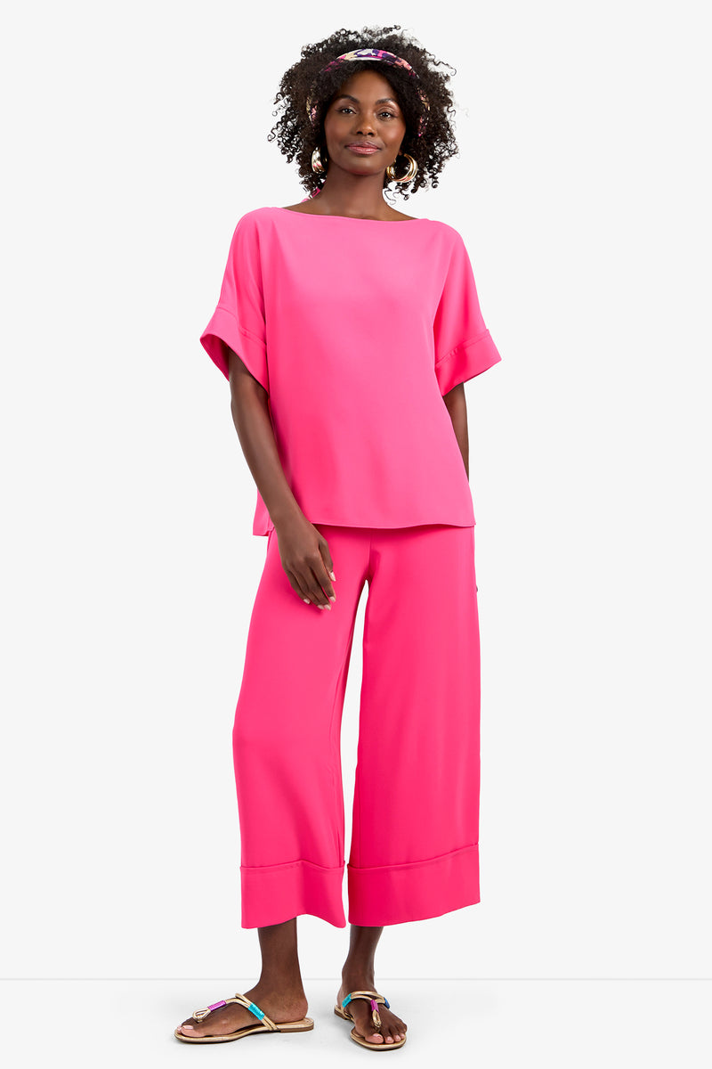 A woman with curly hair and a pink headband smiles, wearing the SOFIE TOP—a loose, bright pink short-sleeve top in soft 100% polyester—paired with matching wide-leg pants and colorful sandals, set against a plain white background.