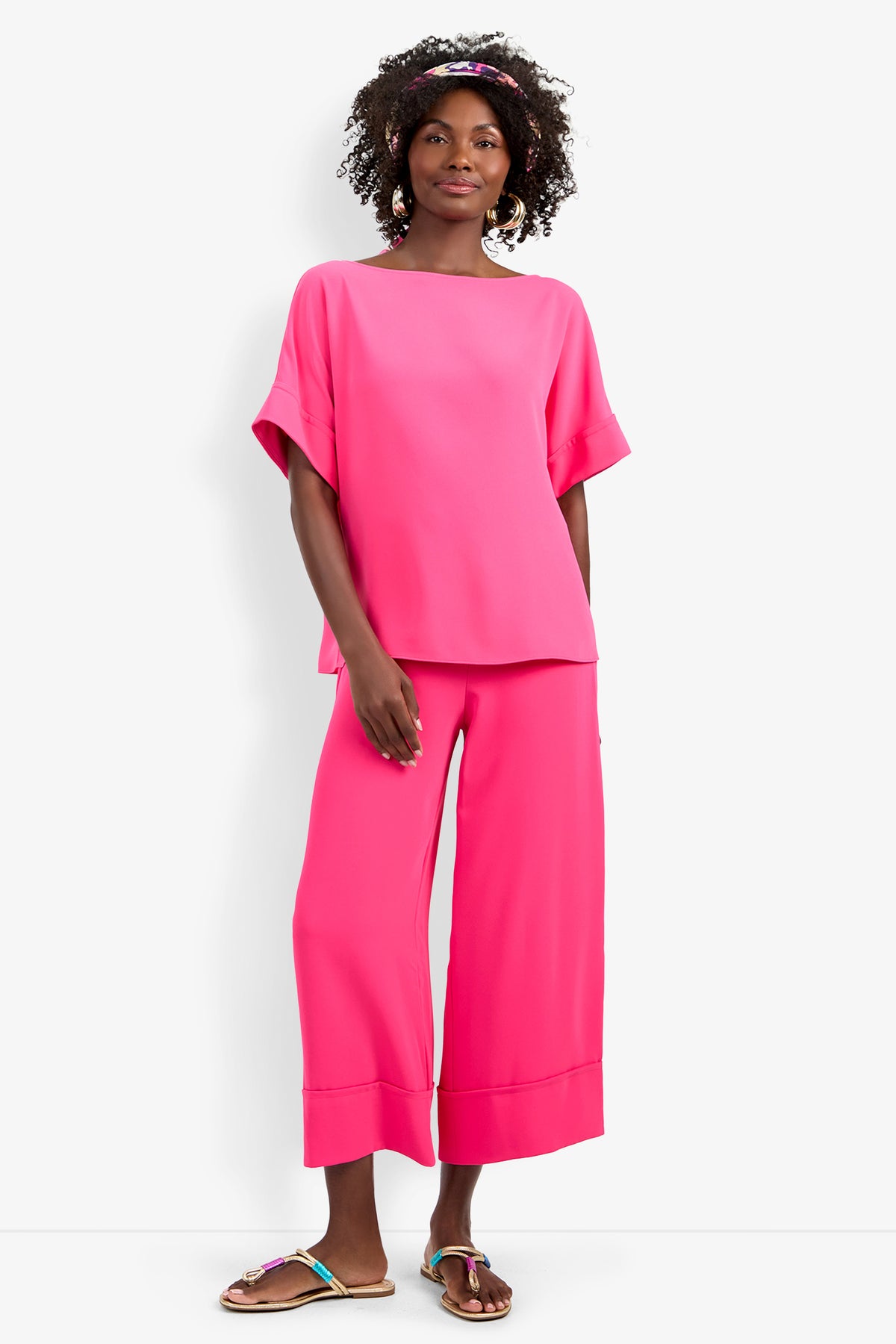 A woman with curly hair and a pink headband stands against a white background wearing the Sofie Top in bright pink with matching wide-leg pants and colorful sandals.
