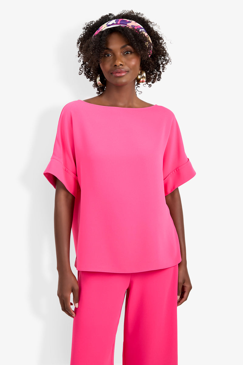 A woman with curly hair, a patterned headband, and gold hoop earrings stands against a white background wearing the bright pink Sofie Top with cuffed short sleeves and matching wide-leg pants.