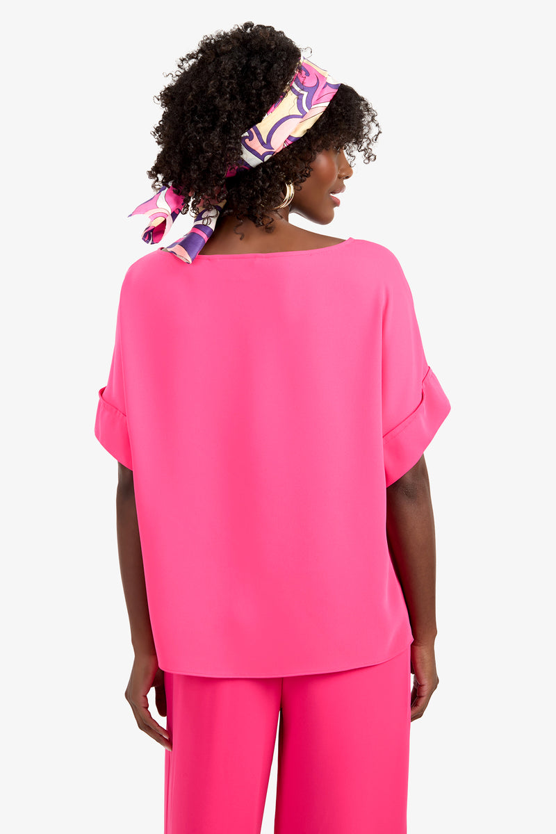 A woman with curly hair, wearing a colorful headscarf, stands with her back to the camera. She is dressed in the bright pink SOFIE TOP and matching pants, both made from soft polyester fabric, against a white background.