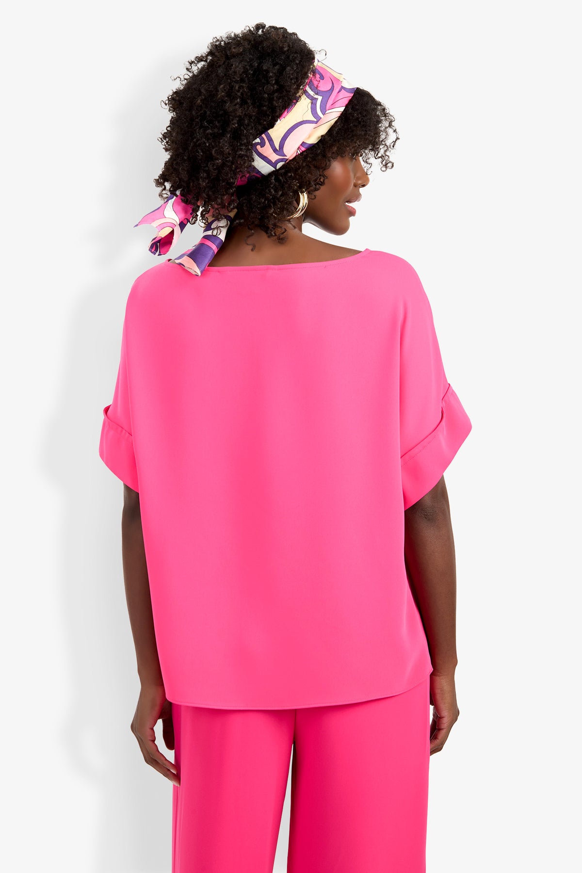 A woman with curly hair, wearing a colorful patterned headscarf and the bright pink Sofie Top with matching pink pants, stands facing away from the camera against a white background.