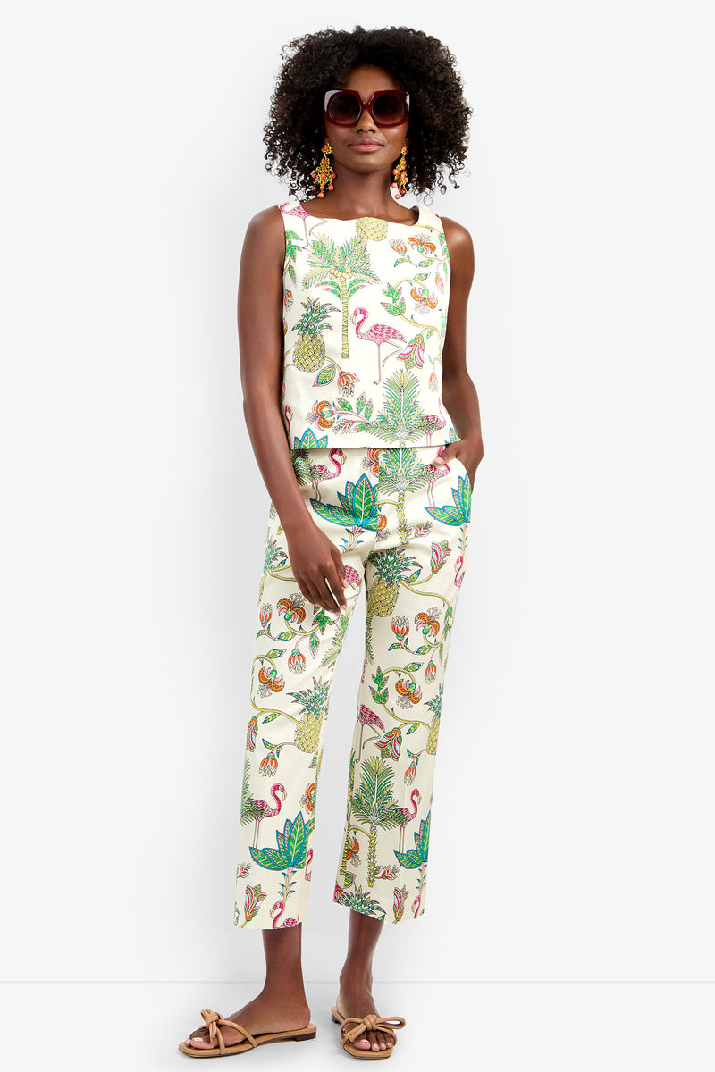 A woman with curly hair wears large sunglasses, statement earrings, and the Venya Top with matching tropical-print pants featuring flamingos, leaves, and pineapples. She stands confidently in tan sandals.