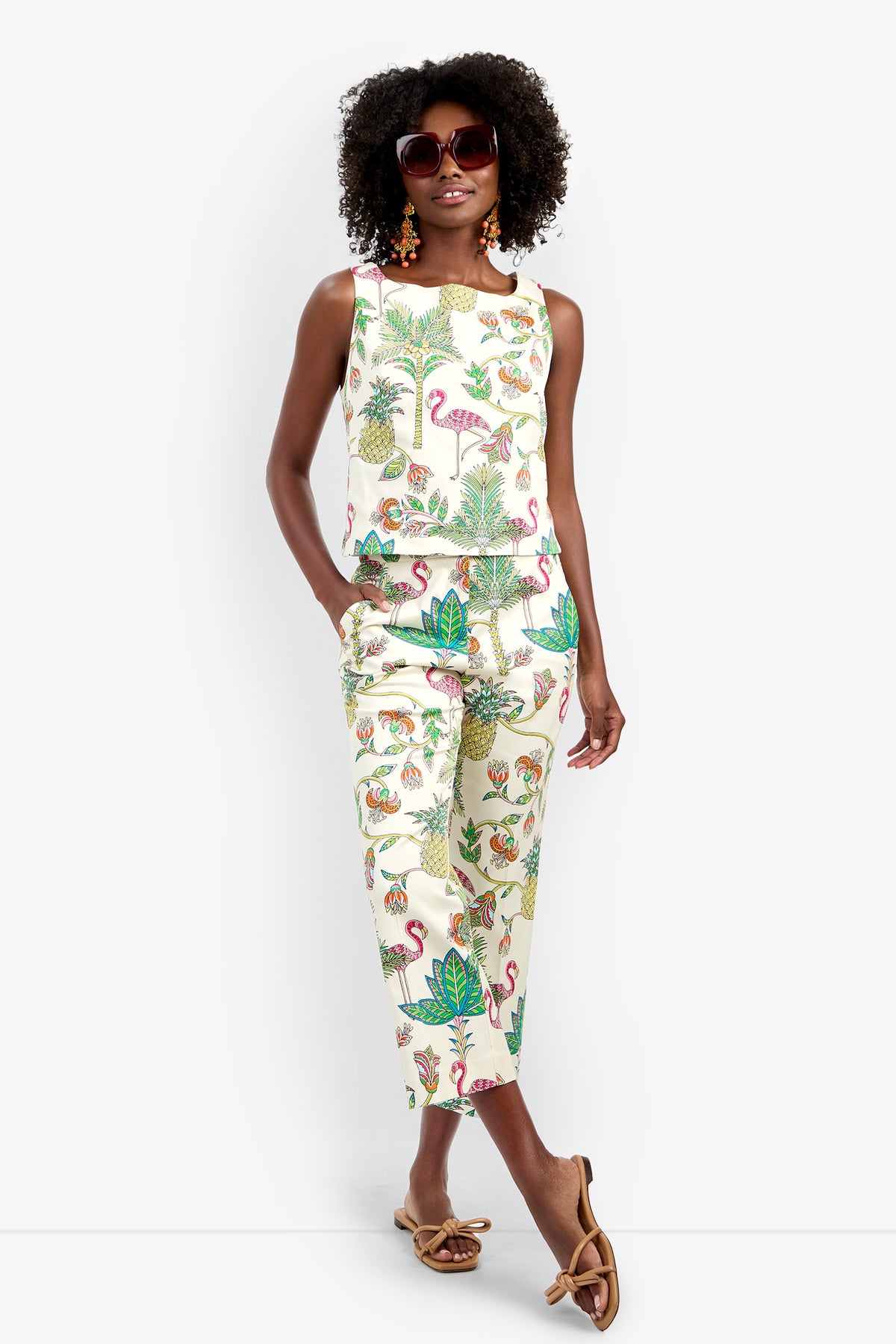 A woman wearing the Venya Top and matching tropical print pants with flamingos and pineapples, paired with sunglasses, earrings, and flat bow sandals, stands against a plain white background.