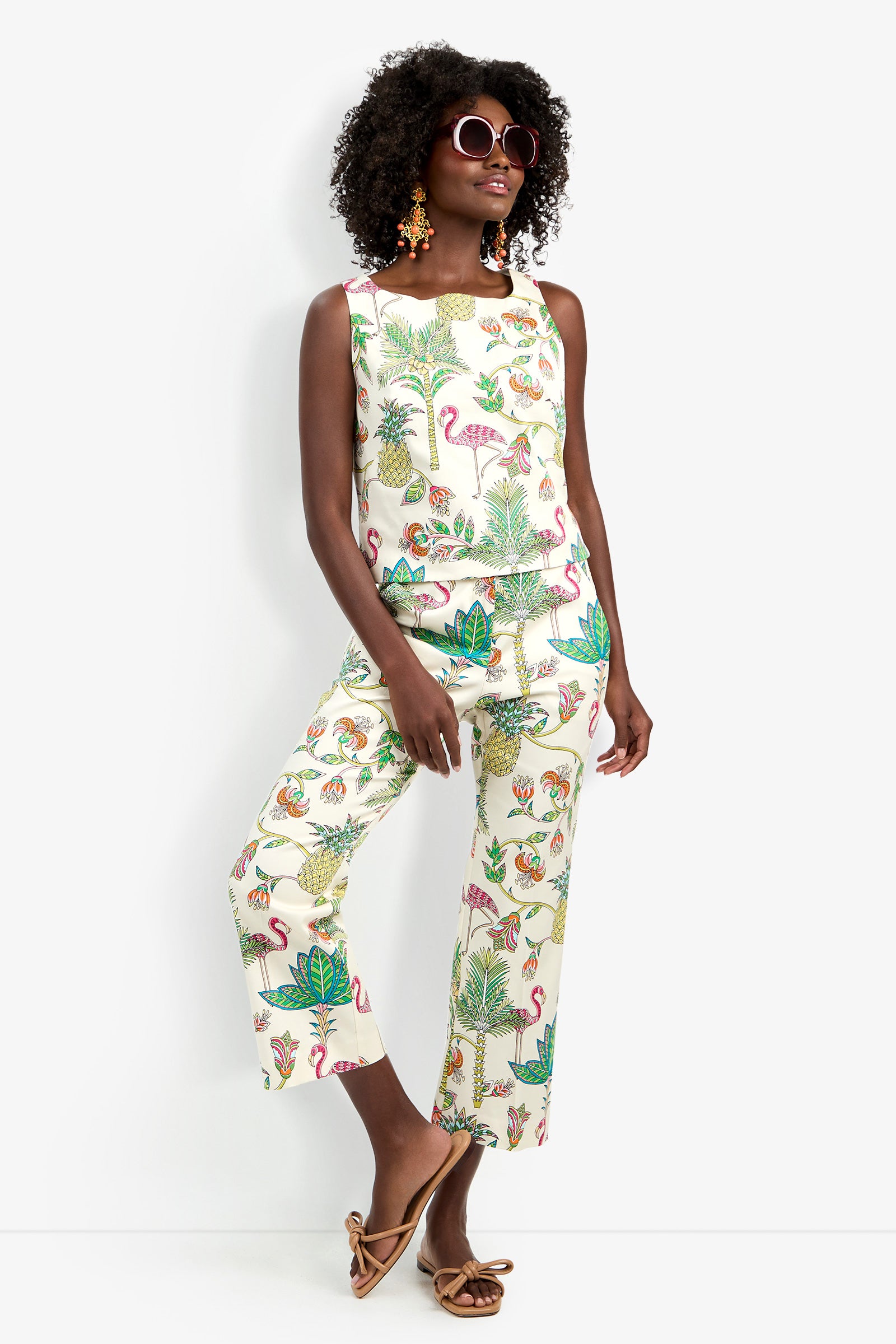 A woman wears the Venya Top—a sleeveless, colorful tropical print with flamingos—paired with matching wide-leg pants, statement earrings, sunglasses, and flat sandals, posing confidently against a white background.