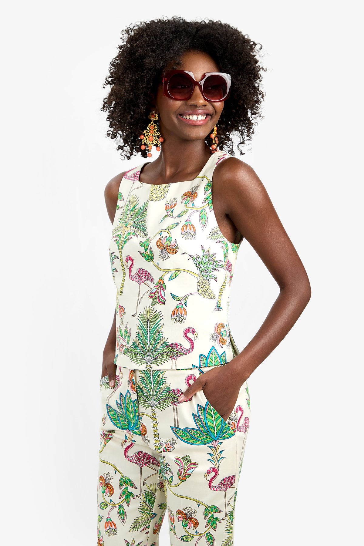 A woman with curly hair smiles, wearing large sunglasses, colorful earrings, and the VENYA 2 TOP—a sleeveless, white cotton-spandex top with a vibrant tropical flamingo and leaf print. Her hands are in her pockets.