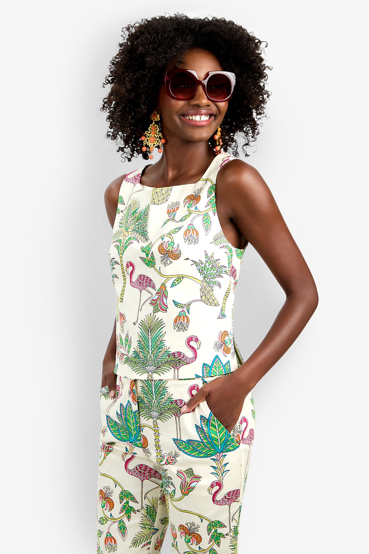A woman with curly hair smiles, hands in pockets, wearing the Venya Top and matching pants—a sleeveless set with a vibrant tropical print featuring flamingos, leaves, and flowers—paired with large sunglasses and colorful earrings.