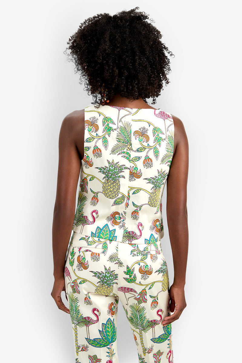 A person with curly hair is seen from behind wearing the Venya Top and matching pants, both featuring colorful tropical prints of pineapples, birds, and leaves on a cream background.
