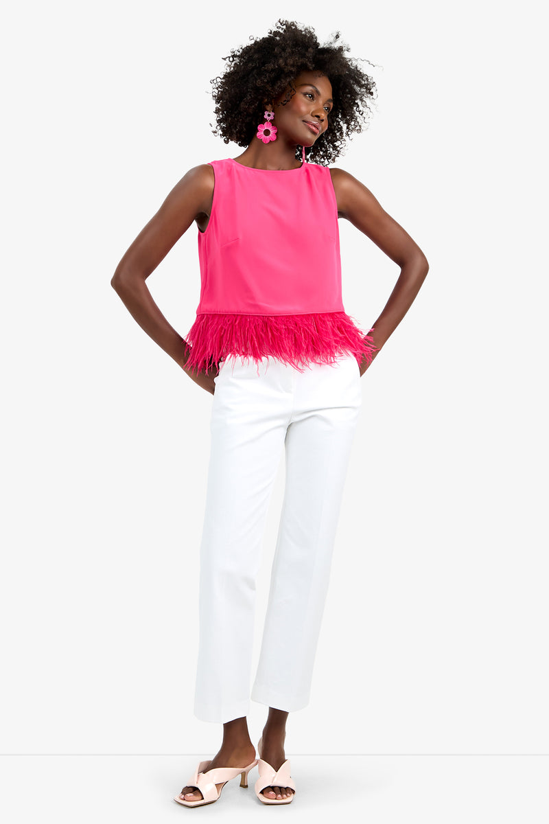 A woman smiles with one hand on her hip, wearing the Rubia Top—a bright pink sleeveless top with feather trim—paired with white wide-leg pants, pink statement earrings, and light pink heeled sandals.