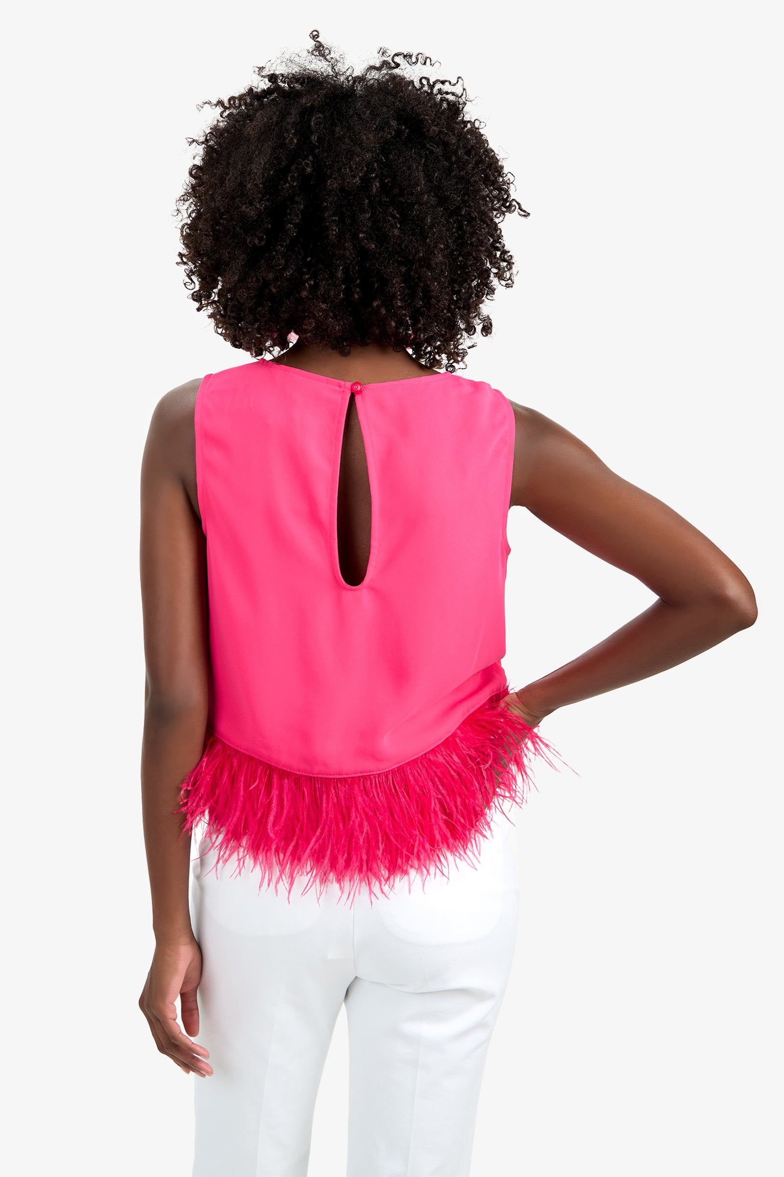 A person with curly hair, seen from behind, wears the Rubia Top—a sleeveless hot pink piece with a keyhole back and feathery hem—paired with white pants.