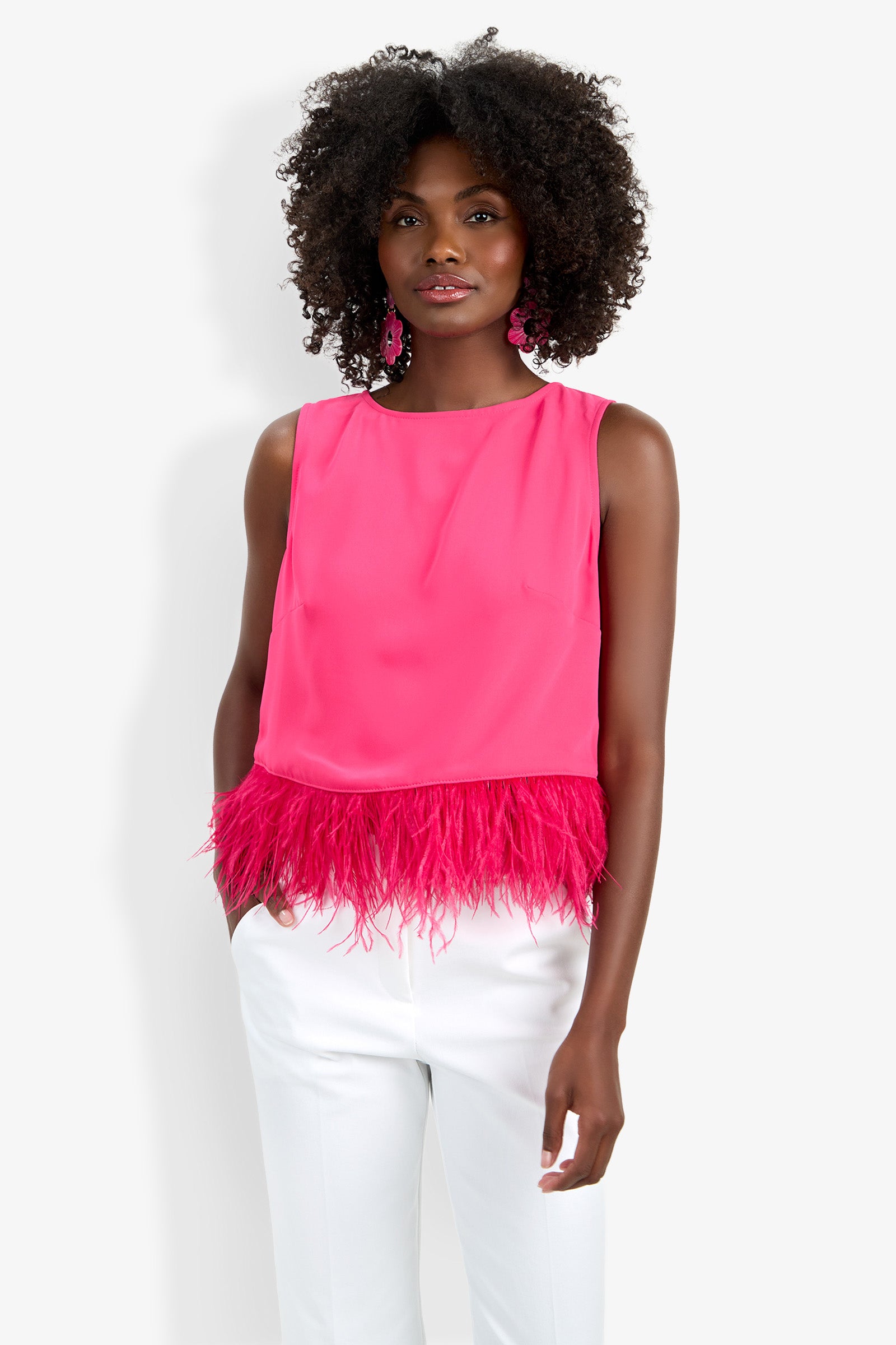 A woman with curly hair wears the Rubia Top—a sleeveless, bright pink blouse with feather trim at the hem—paired with white pants. She stands against a plain white background.