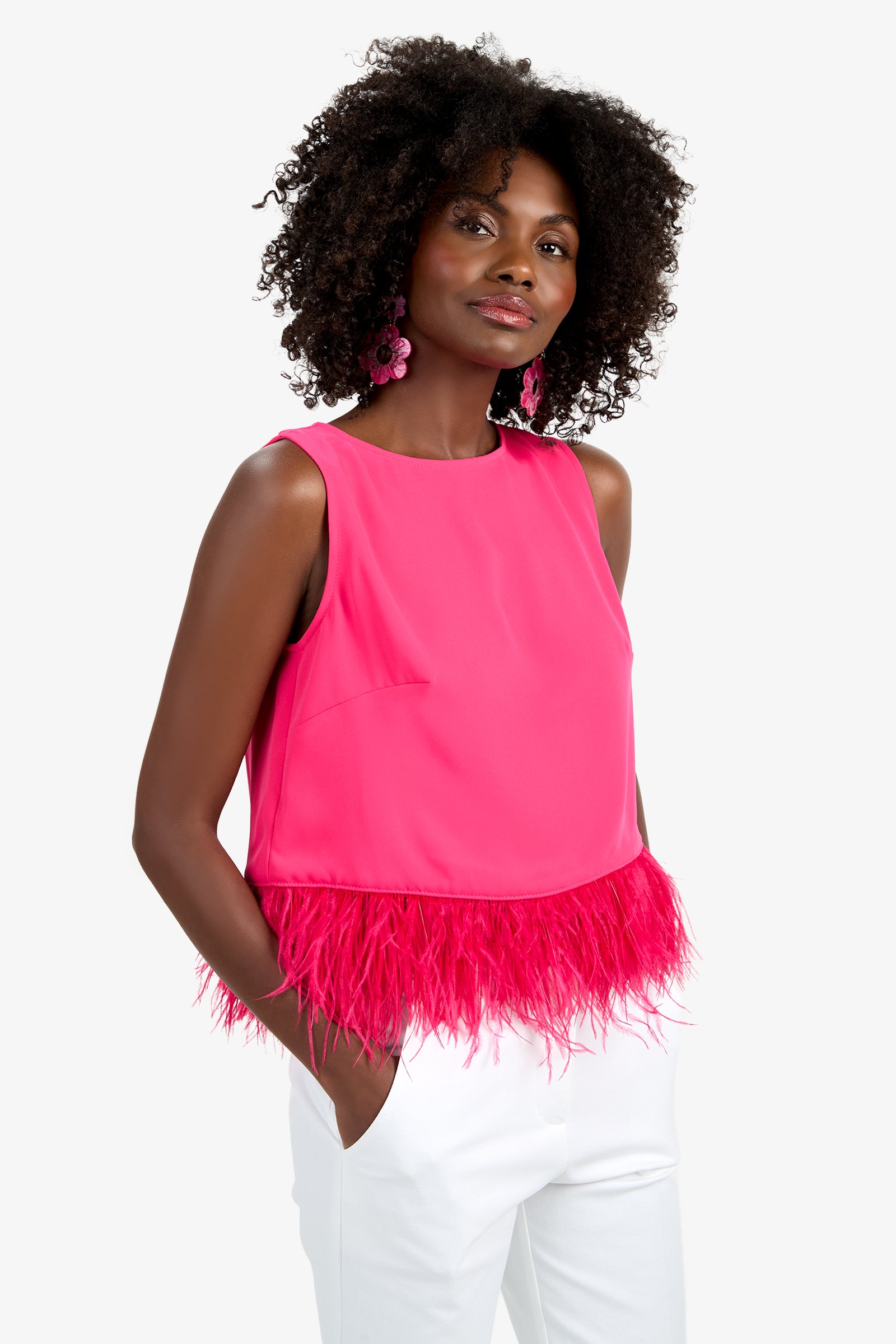A woman with curly hair wears the Rubia Top—a bright pink sleeveless top with a feathered hem—paired with white pants and statement pink earrings. She stands with one hand in her pocket, smiling slightly.