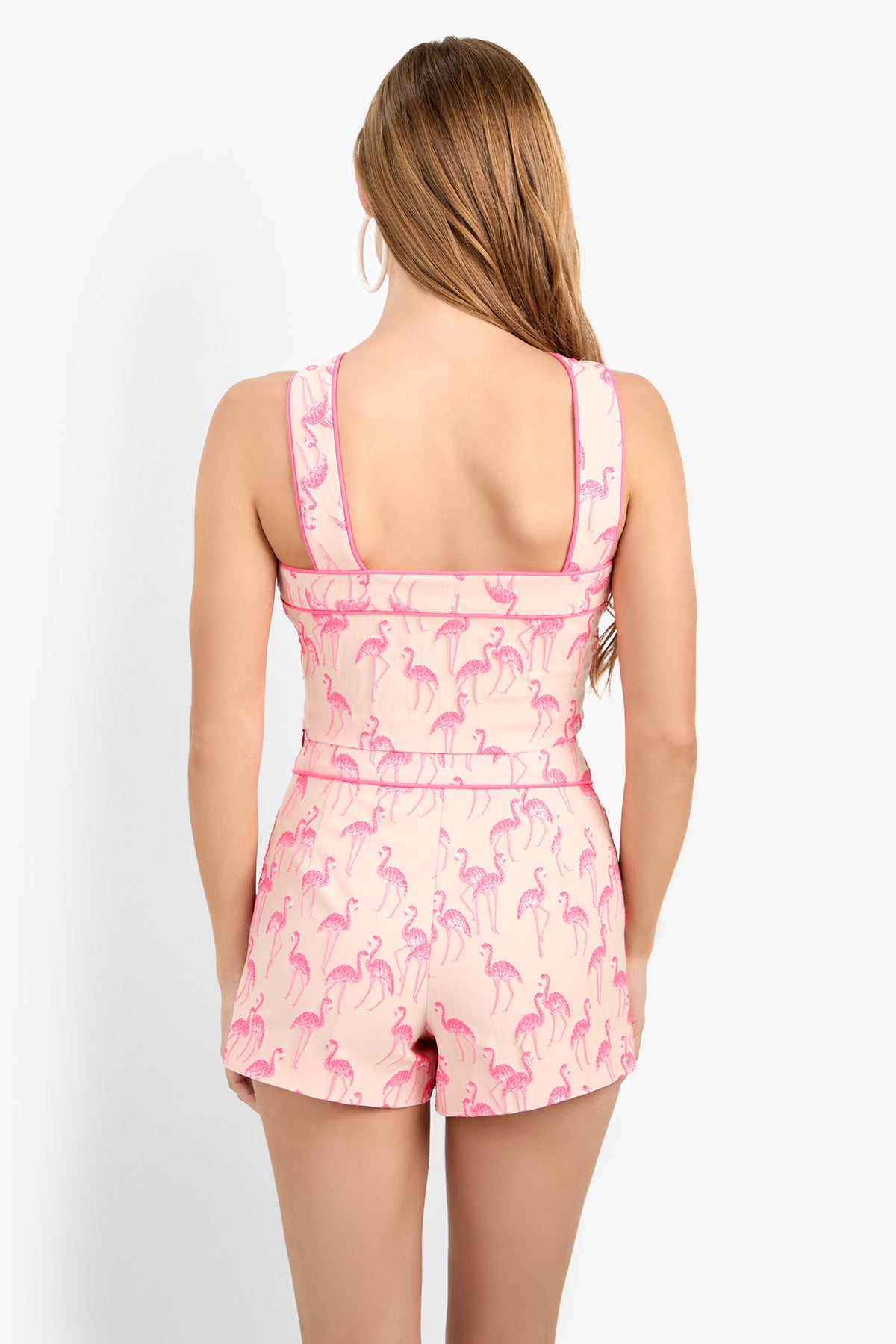 A woman with long light brown hair stands with her back to the camera, wearing the sleeveless PIPED WANONA TOP, crafted from soft polyester and featuring a playful flamingo print.
