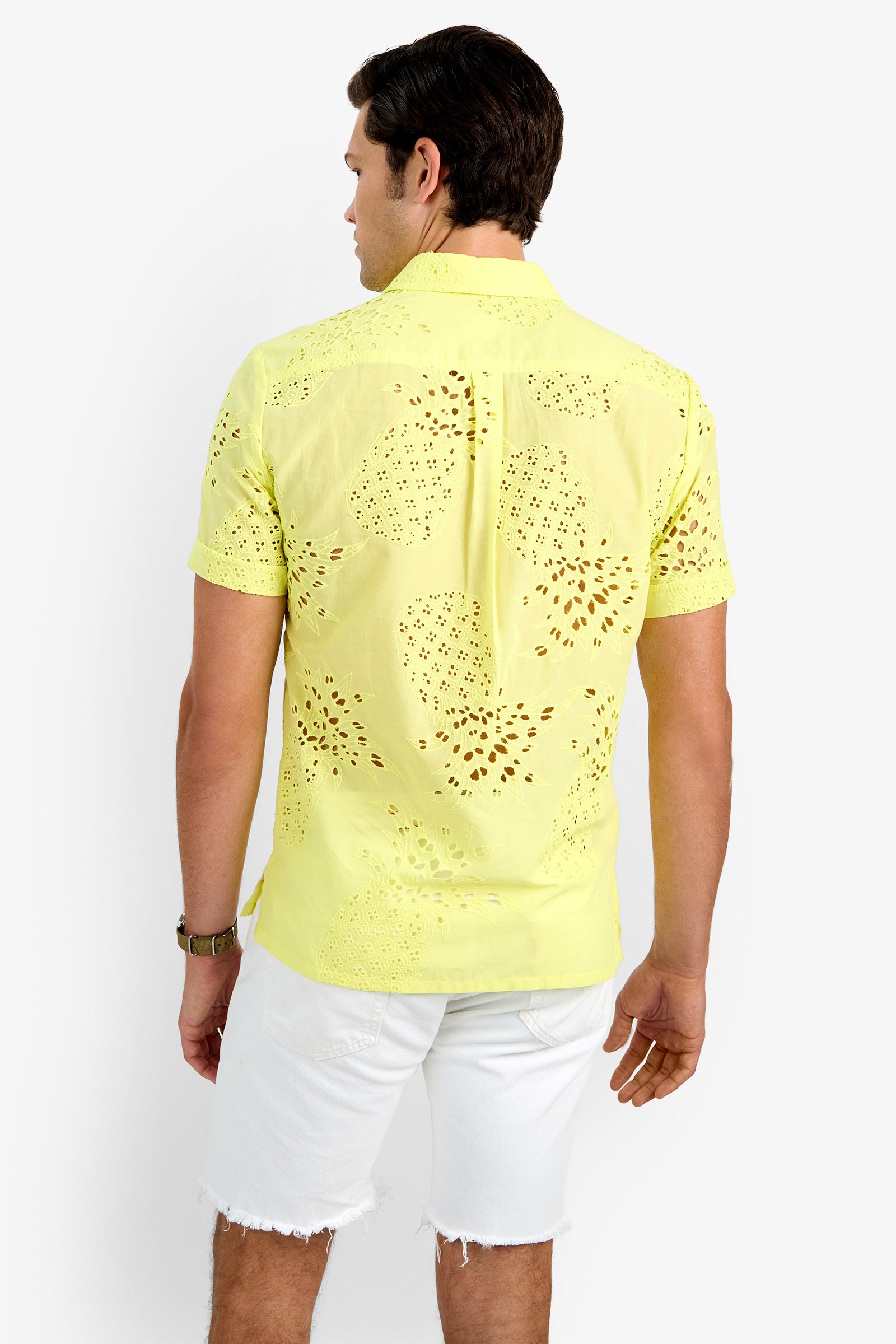 A man with short dark hair and a watch stands with his back to the camera, wearing the BRANSON SHIRT in light yellow (100% cotton, perforated) and white frayed-hem shorts against a plain white background.