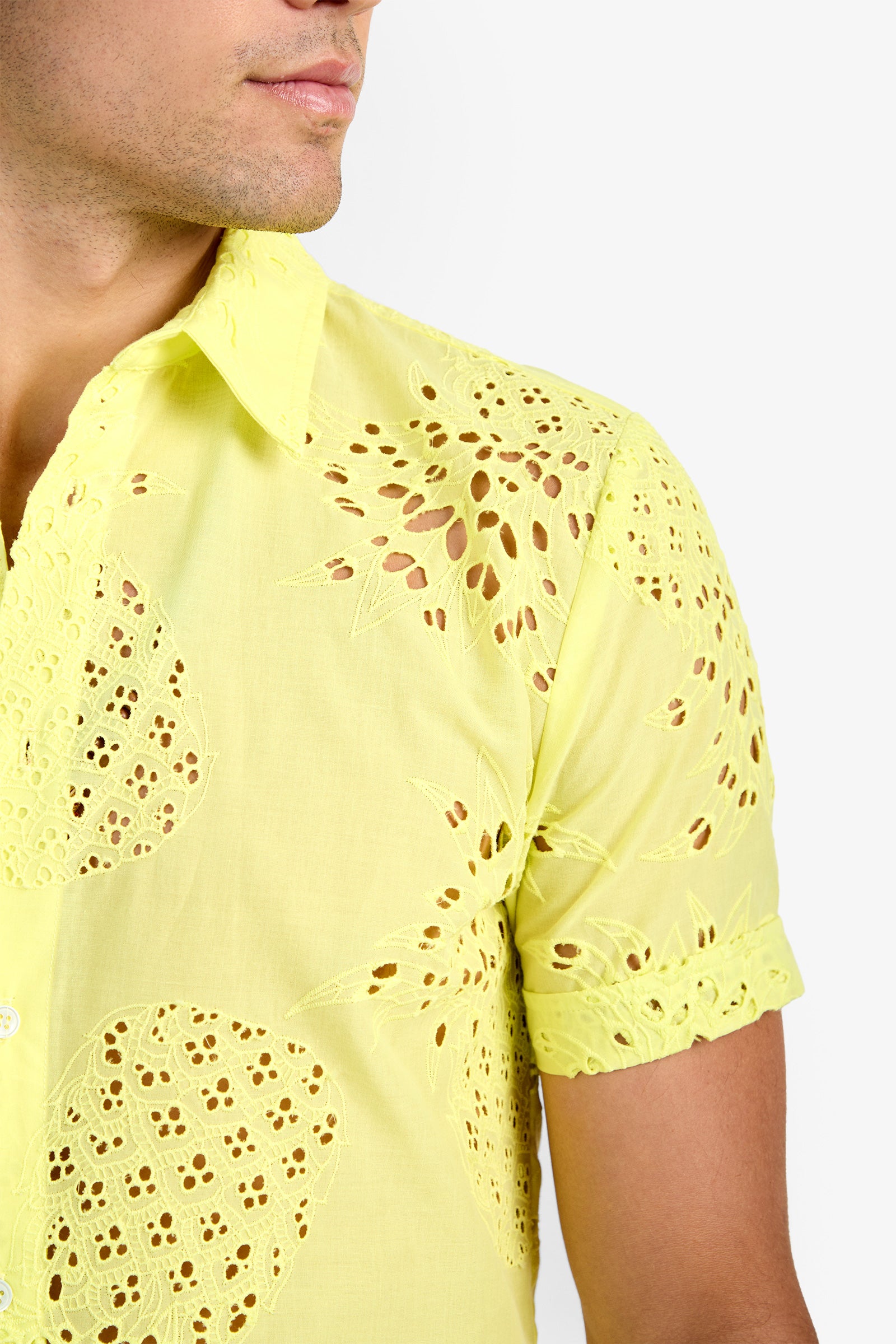 A close-up of a person wearing the BRANSON SHIRT, a light yellow, short-sleeve, 100% cotton button-up with intricate cut-out and embroidered circular and floral patterns. The person's face is partially visible.