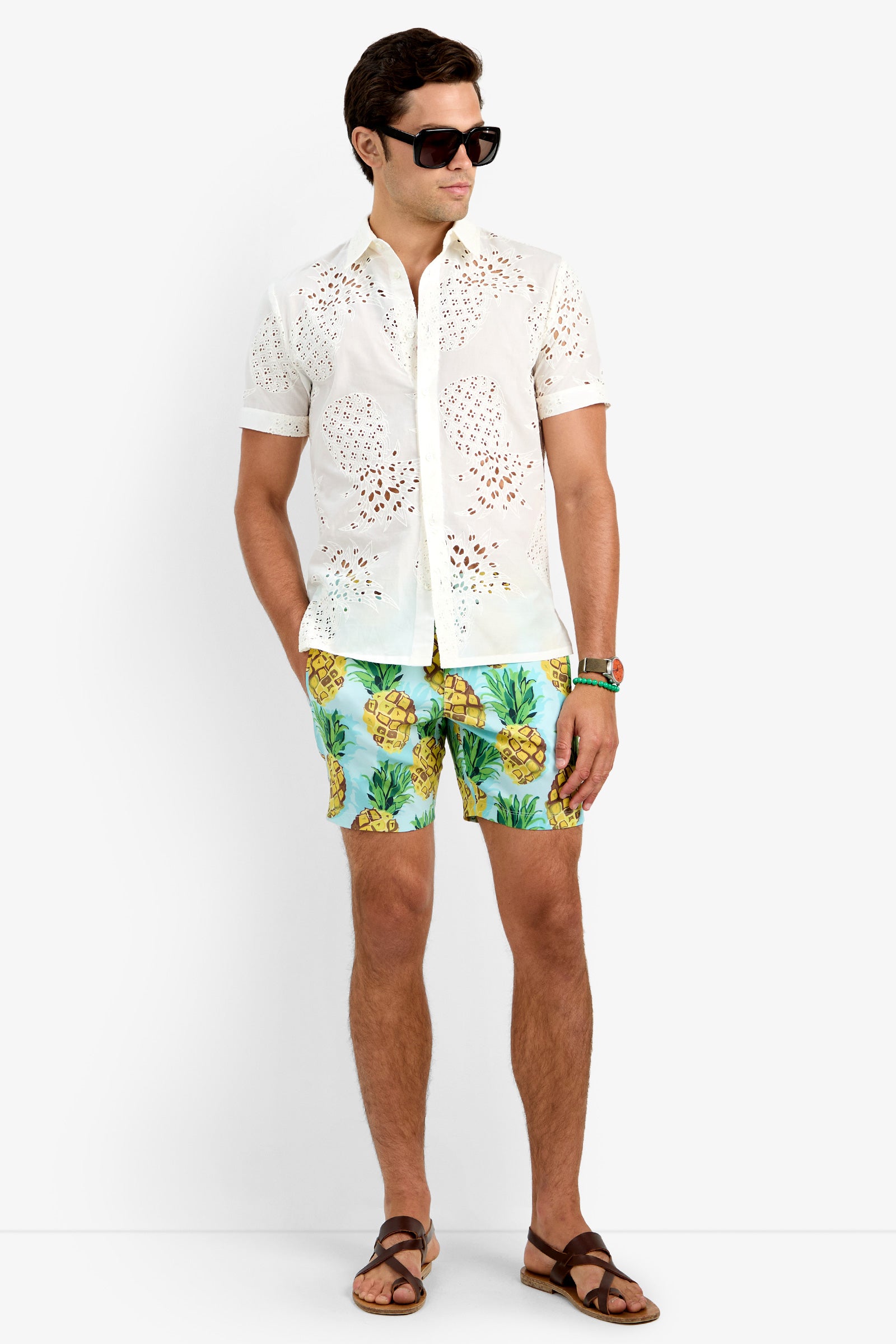 A man wearing black sunglasses, the BRANSON SHIRT (white, short-sleeve, 100% cotton eyelet), colorful pineapple-print shorts, brown sandals, and a watch stands against a plain white background.