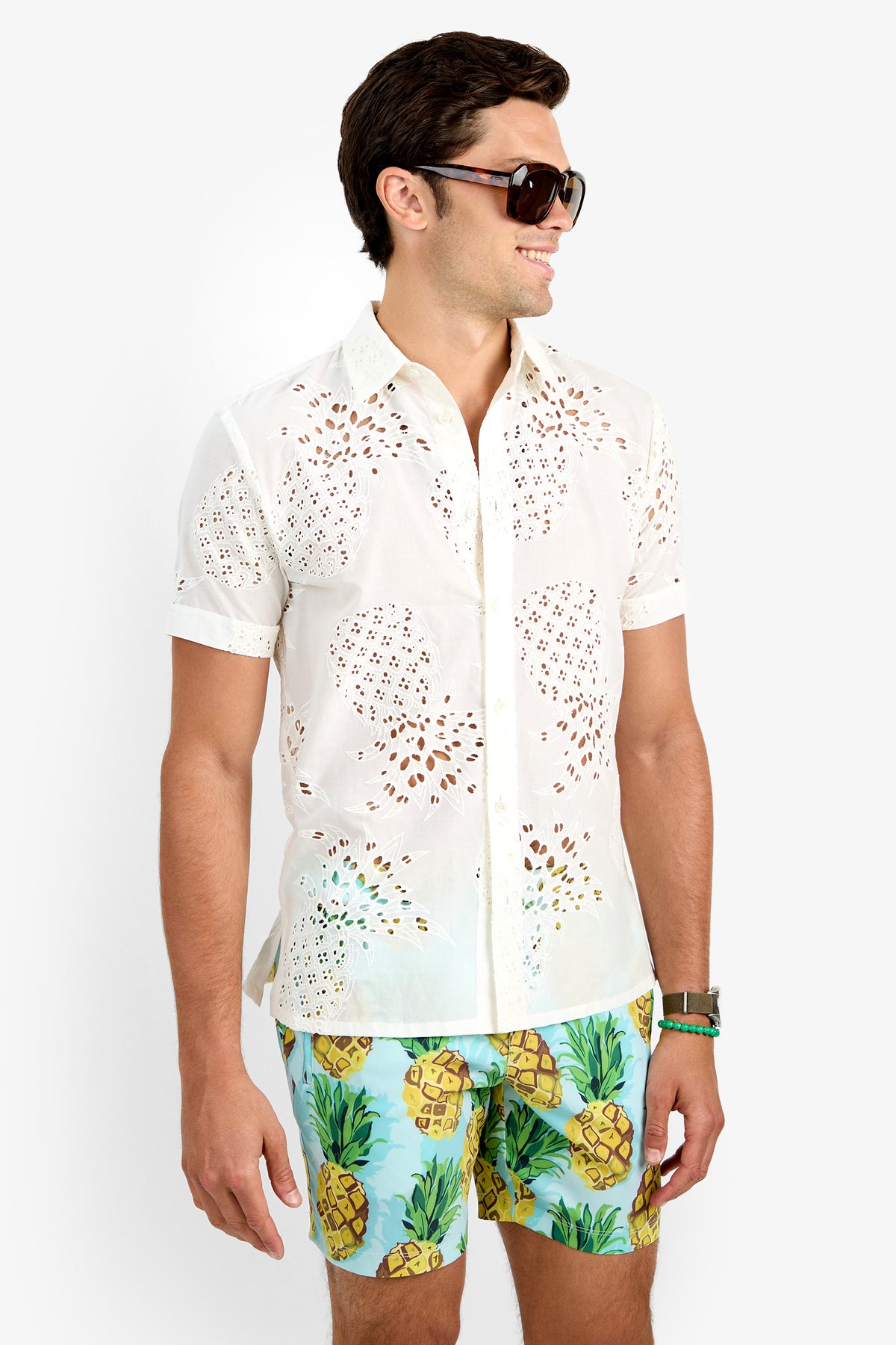 A man smiles and looks to the side, wearing large sunglasses, colorful pineapple print swim shorts, and the BRANSON SHIRT—a short-sleeve white eyelet shirt made from 100% cotton—against a plain white background.