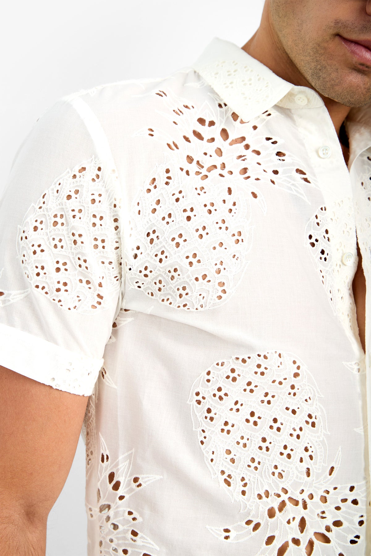 A close-up of a person wearing the BRANSON SHIRT—a white, short-sleeved, button-up shirt made from 100% cotton with intricate circular lace-like cutouts—shows only part of their face and upper body.