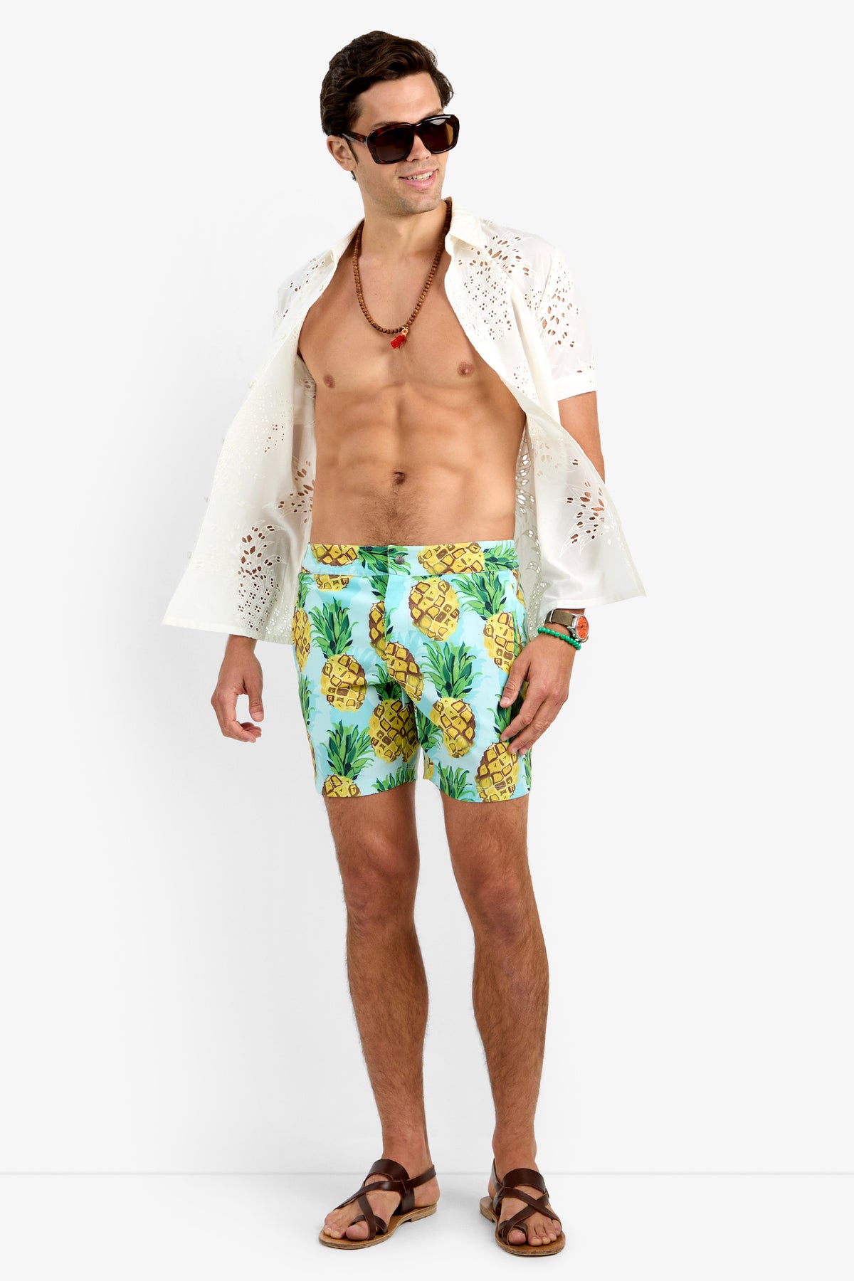 A man wearing sunglasses, the BRANSON SHIRT (white, 100% cotton, openwork), blue pineapple-print swim trunks, brown sandals, and a beaded necklace stands smiling against a plain white background.