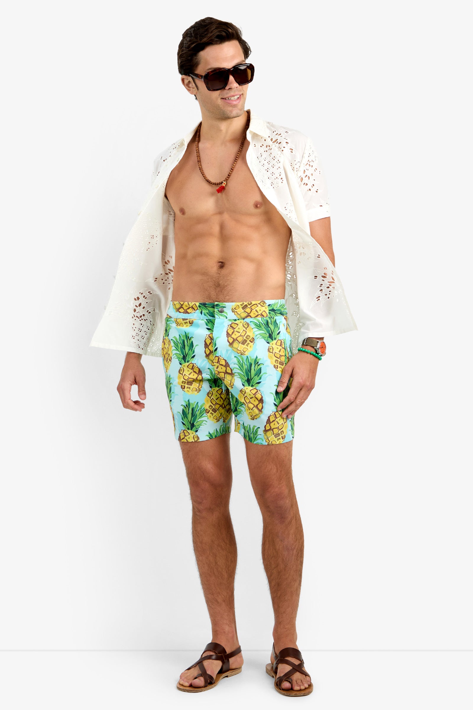A man wearing sunglasses, the BRANSON SHIRT (white, 100% cotton, openwork), blue pineapple-print swim trunks, brown sandals, and a beaded necklace stands smiling against a plain white background.