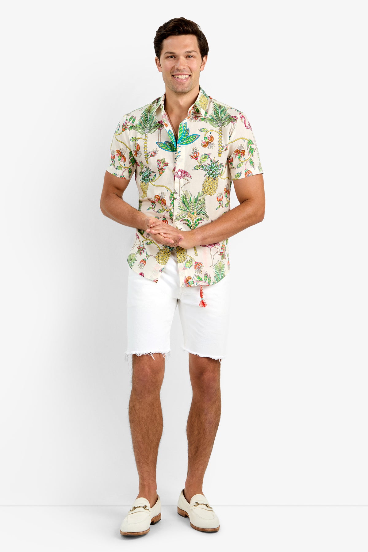 A man smiles in the SLIM JIM SHIRT, a tropical print cotton short-sleeve, paired with white frayed-hem shorts and cream loafers without socks. He stands against a plain white background, hands loosely clasped.