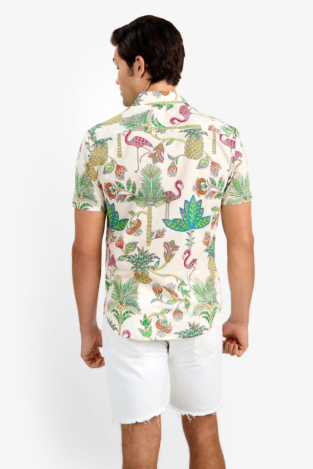 A man faces away, wearing the SLIM JIM SHIRT—a short-sleeve, cream 100% cotton shirt with a vibrant tropical flamingo and plant print—paired with white frayed-hem shorts.