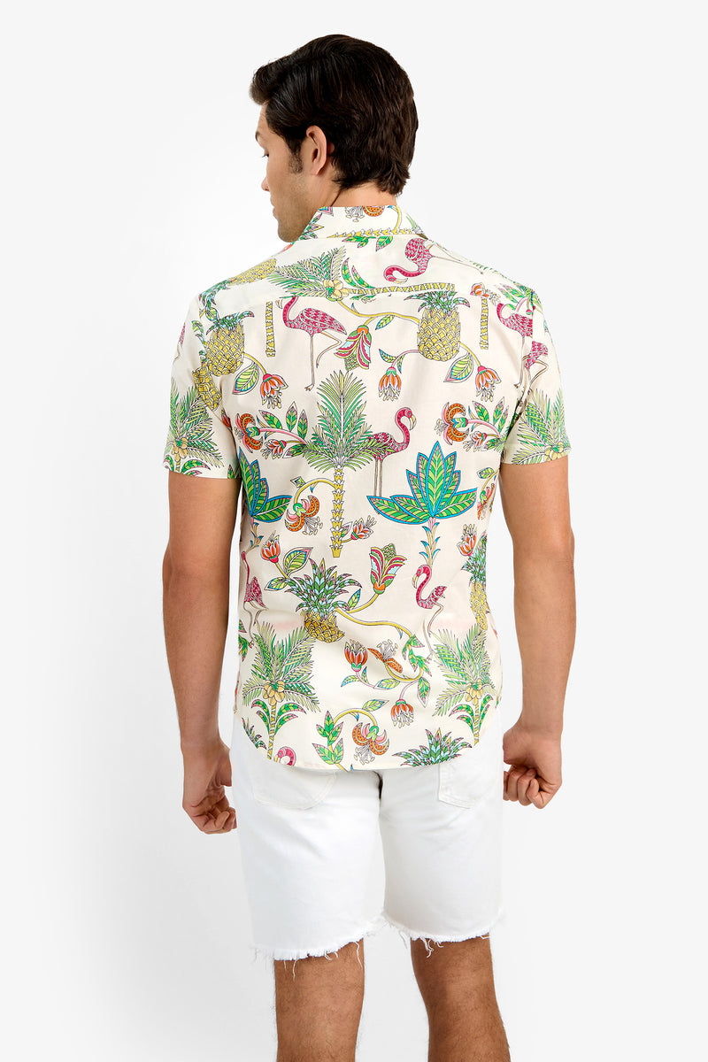 A man faces away, wearing the SLIM JIM SHIRT—a short-sleeve, cream 100% cotton shirt with a vibrant tropical flamingo and plant print—paired with white frayed-hem shorts.