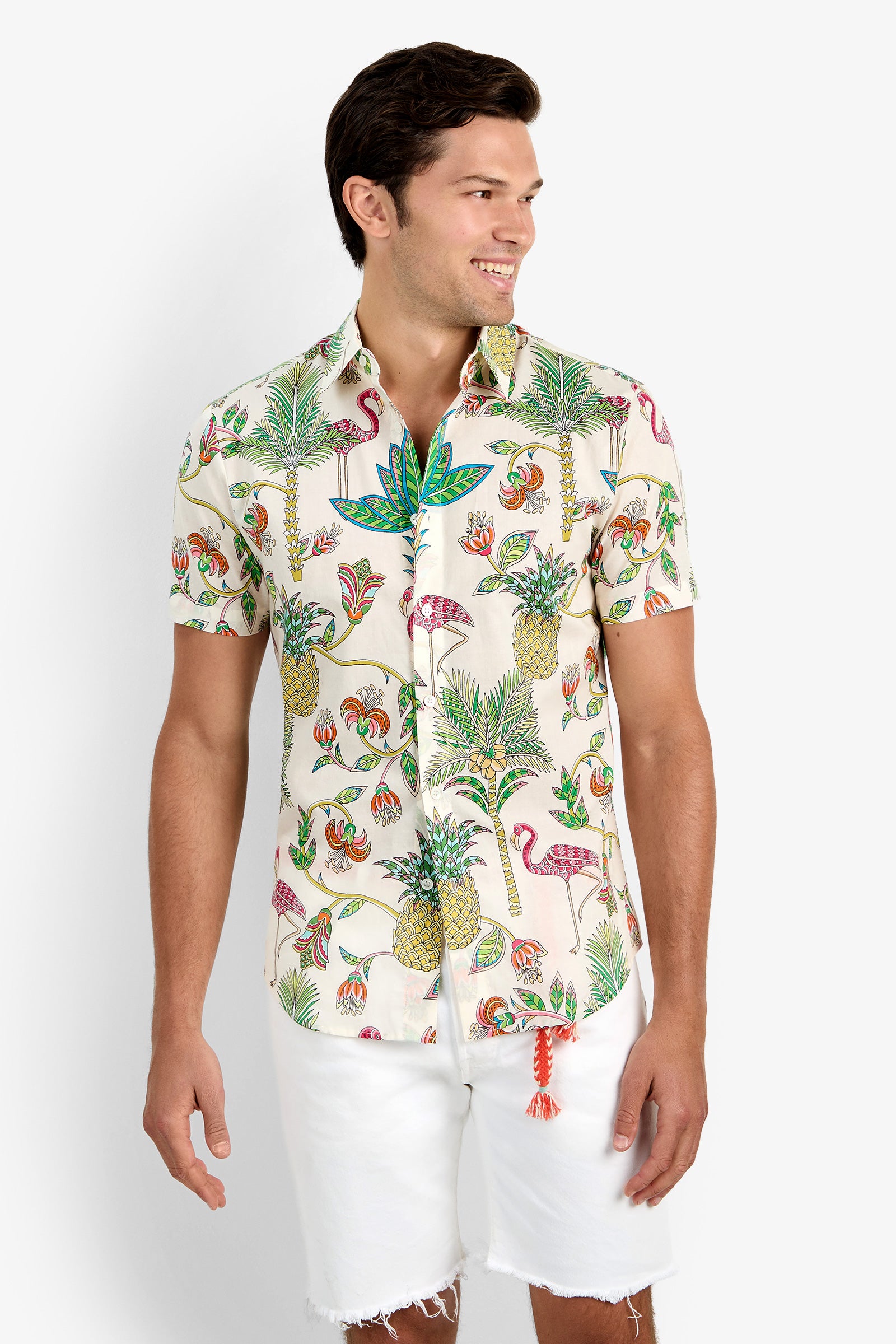 A man smiles and looks to the side against a plain background, wearing the SLIM JIM SHIRT—a short-sleeve, 100% cotton button-up with a colorful tropical pineapple, palm tree, and bird print—paired with white shorts.