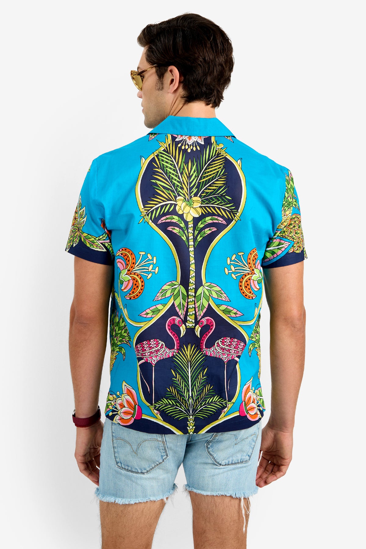 A man in sunglasses and the JOSUE CAMP SHIRT—a bright blue, 100% cotton short-sleeve with colorful tropical prints—paired with light blue distressed denim shorts stands facing away from the camera against a plain background.