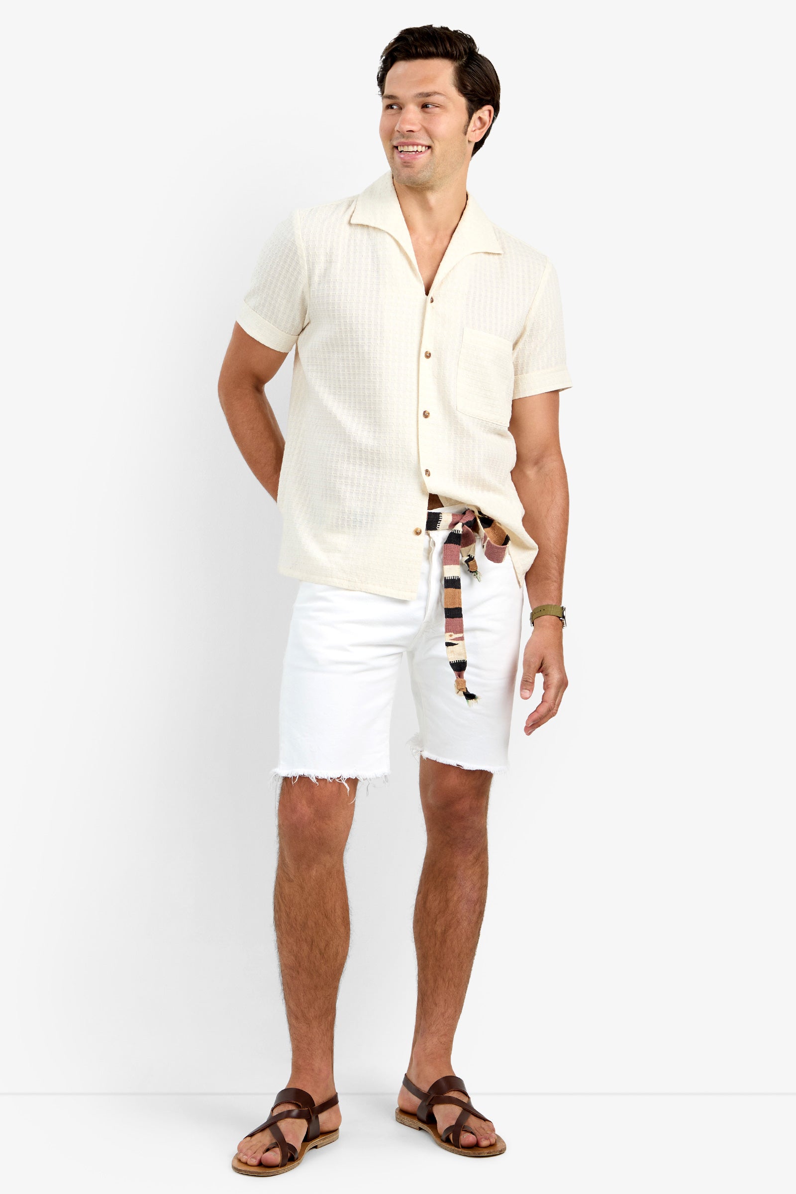 A man with short dark hair smiles against a light background, wearing THEO CAMP SHIRT in ECRU—a soft cotton short-sleeve button-up—paired with white frayed hem shorts, a striped belt, and brown sandals.