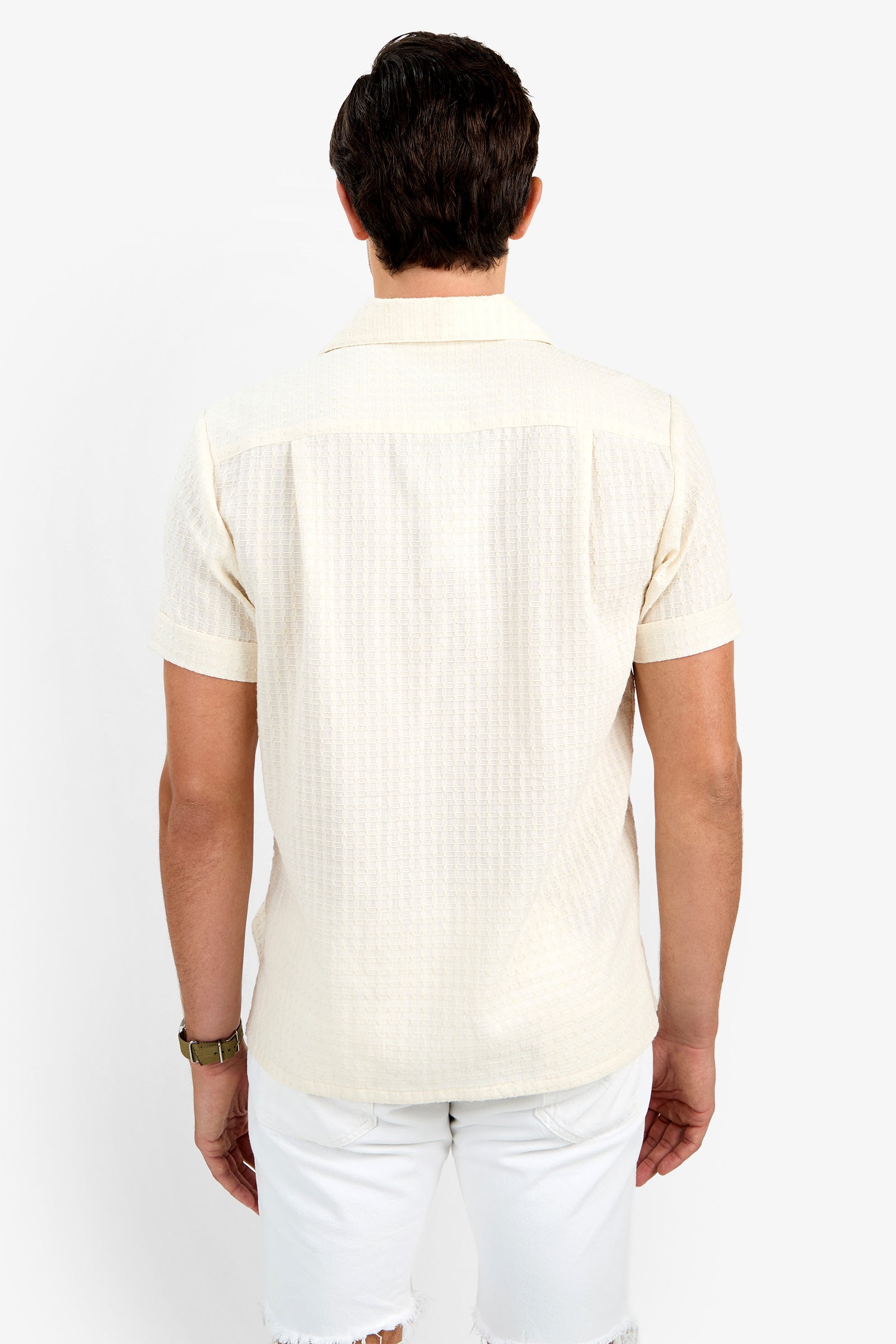 A man with short dark hair is seen from behind wearing the THEO CAMP SHIRT, a light beige 100% cotton short-sleeve shirt, paired with white shorts against a plain white background.