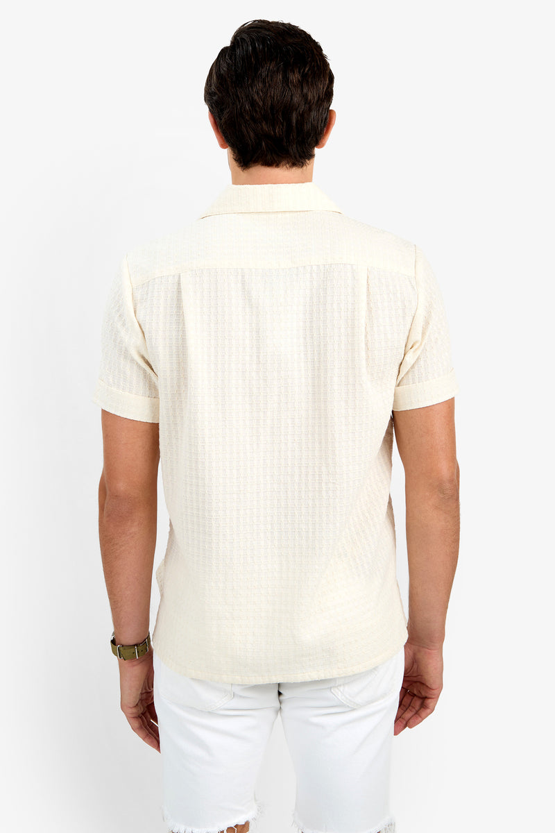 A man with short dark hair is seen from behind wearing the THEO CAMP SHIRT, a light beige 100% cotton short-sleeve shirt, paired with white shorts against a plain white background.