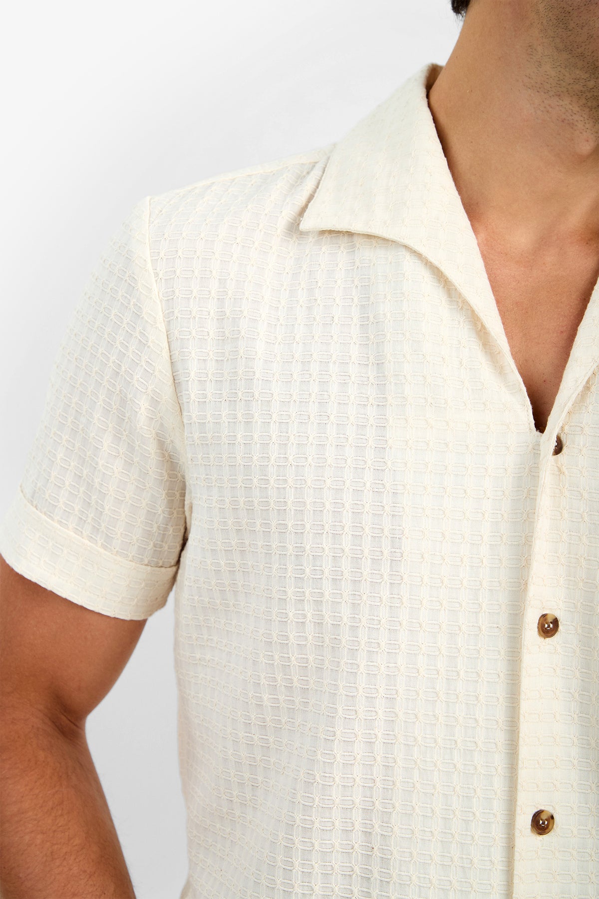 A man wears the THEO CAMP SHIRT— a short-sleeve, cream textured 100% cotton shirt with a camp collar and brown buttons—shown from shoulders to mid-torso against a plain background.