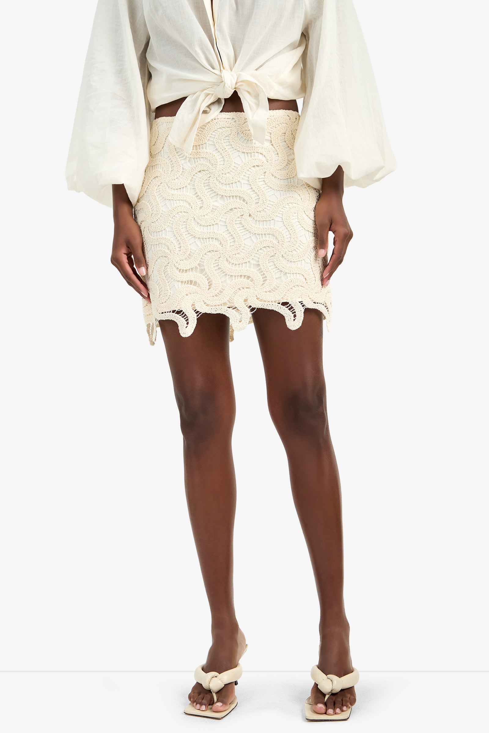 Wearing the RICO SCALLOP SKIRT—a cream, 100% cotton crocheted mini with a wavy pattern—paired with a long-sleeve waist-tied blouse and cream decorative strap sandals, standing against a white background.