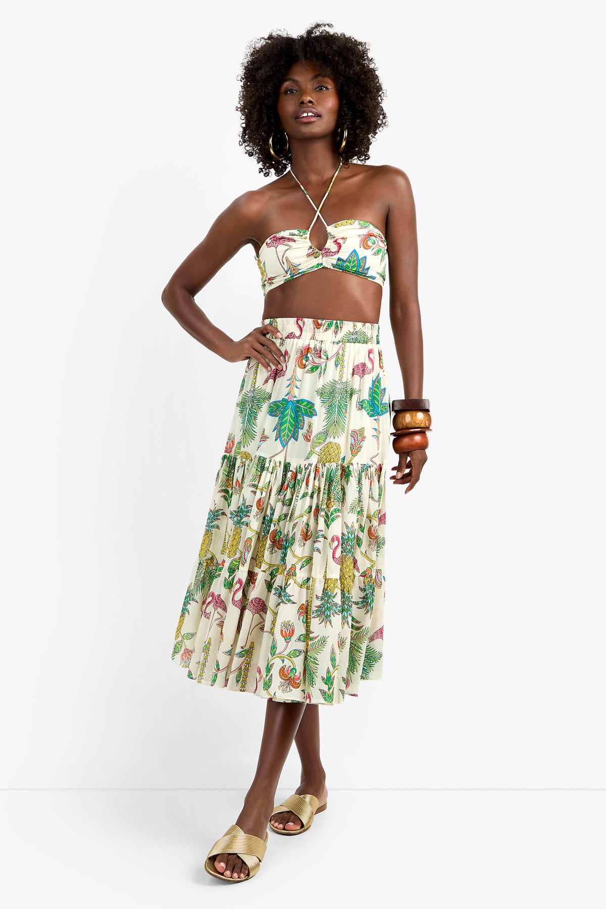 Woman in MARSAY SKIRT, a 100% cotton tiered midi with colorful tropical print, paired with strapless crop top, tan slide sandals, wooden bangles, and hoop earrings against a white background.