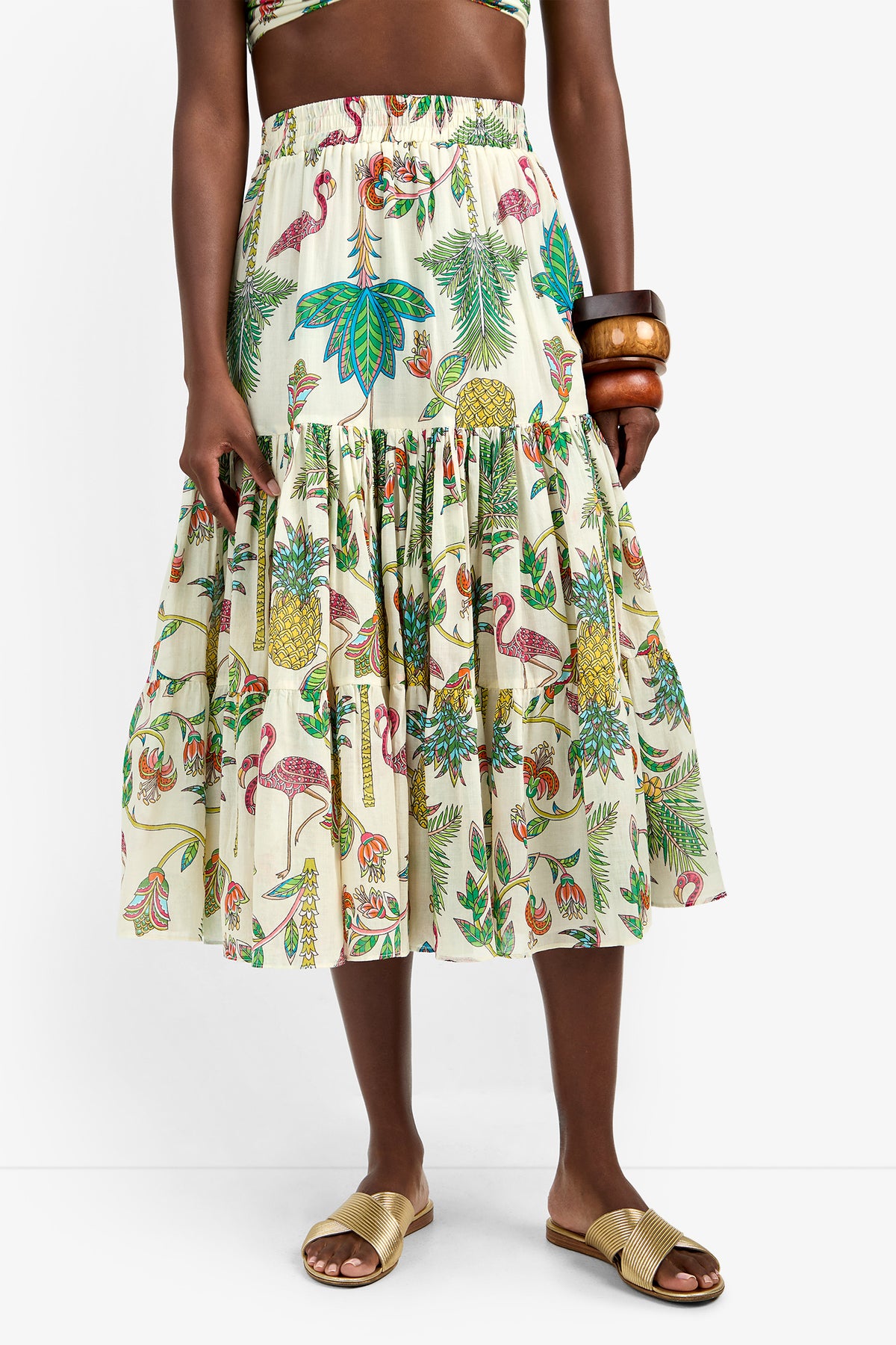 A person wearing the MARSAY SKIRT, a colorful tropical-print 100% cotton midi skirt with leaves, birds, and pineapples, paired with tan slide sandals and chunky wooden bangles. The background is plain white.