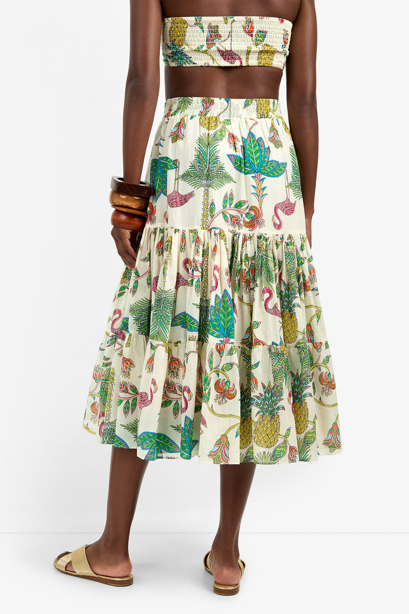 A woman stands against a plain white background, wearing the MARSAY SKIRT—a colorful, tropical-print 100% cotton skirt with a matching strapless top—styled with large bangles and tan slide sandals.
