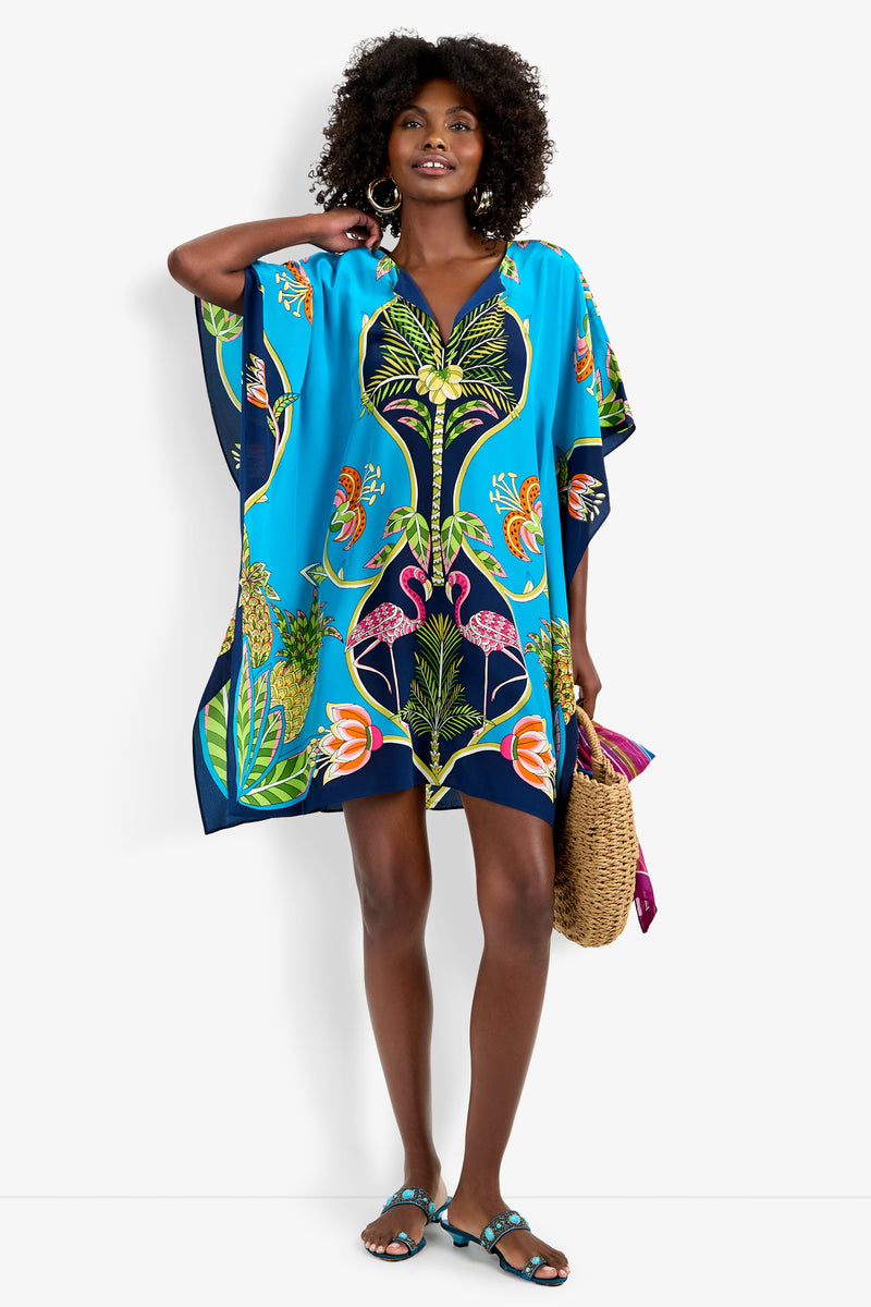 A woman with curly hair smiles, touching her head, while wearing the Theodora Caftan Dress featuring a bright blue tropical print with colorful birds and plants. She carries a woven straw bag and wears sandals.
