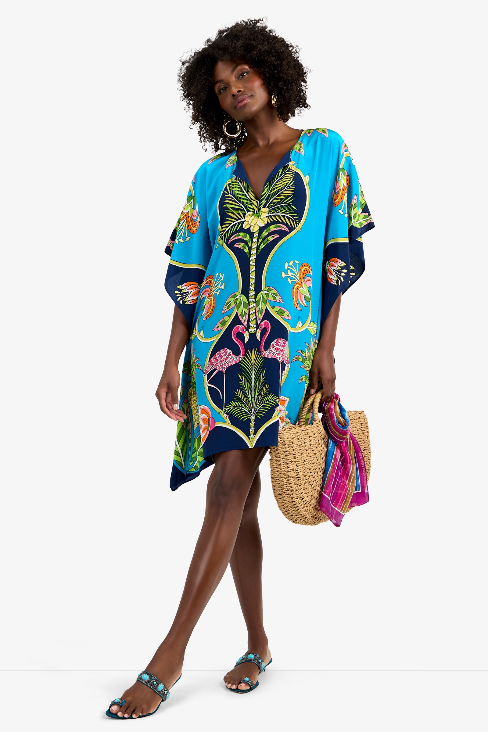 A woman with curly hair and large hoop earrings wears the THEODORA DRESS, a vibrant blue silk tropical print dress with flamingos. She carries a straw tote bag with a pink scarf and wears blue sandals.