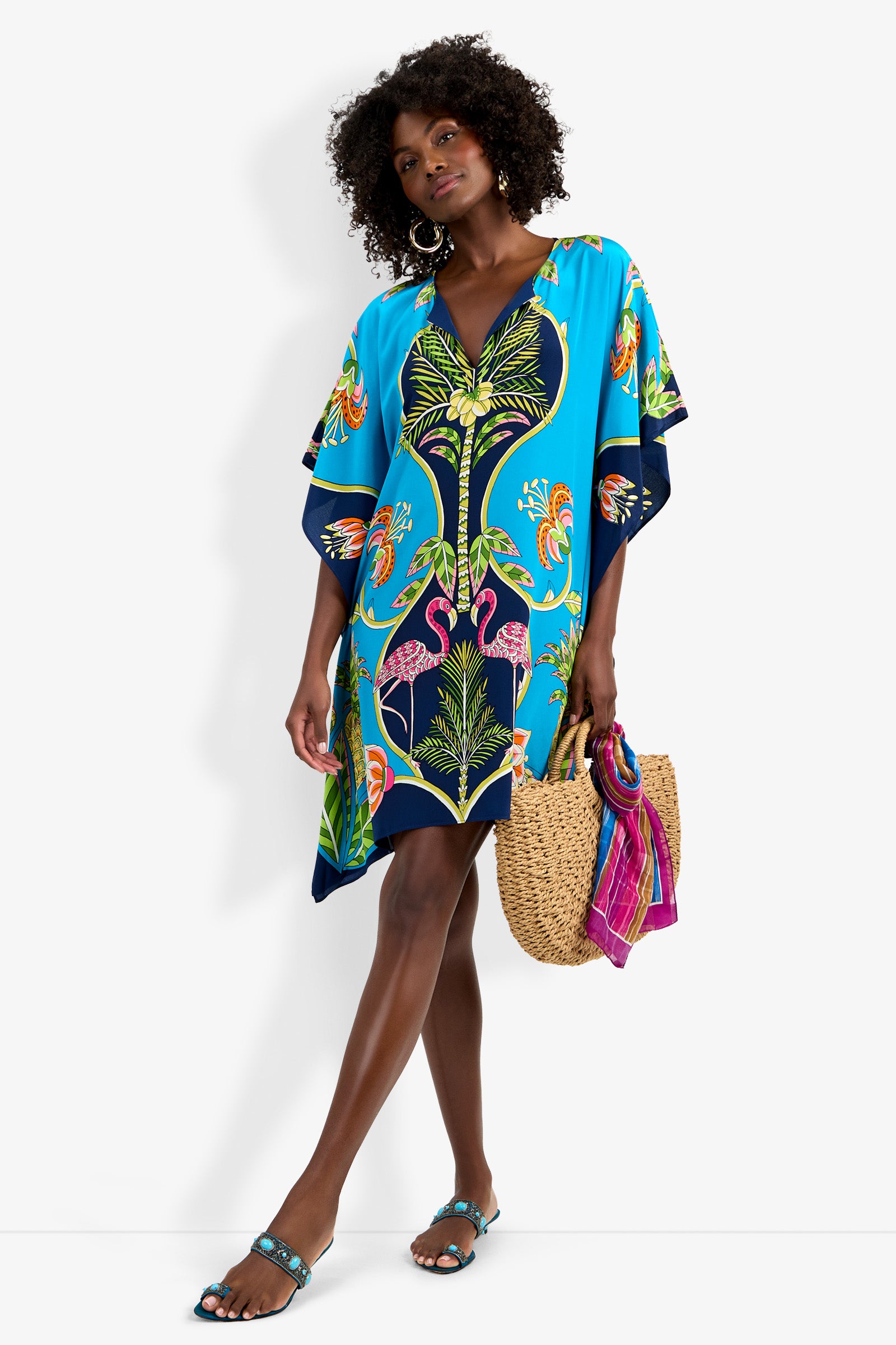 Woman in Theodora Caftan Dress with blue tropical print and pink flamingos, holding a straw bag with a pink scarf, wearing blue sandals and hoop earrings, standing against a white background.
