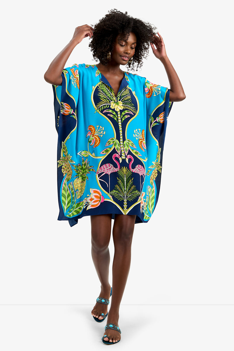 A woman wears the THEODORA DRESS, a bright blue, loose-fitting pure silk kaftan with tropical prints of flamingos, pineapples, and flowers. She raises her arms and completes the look with matching blue sandals.