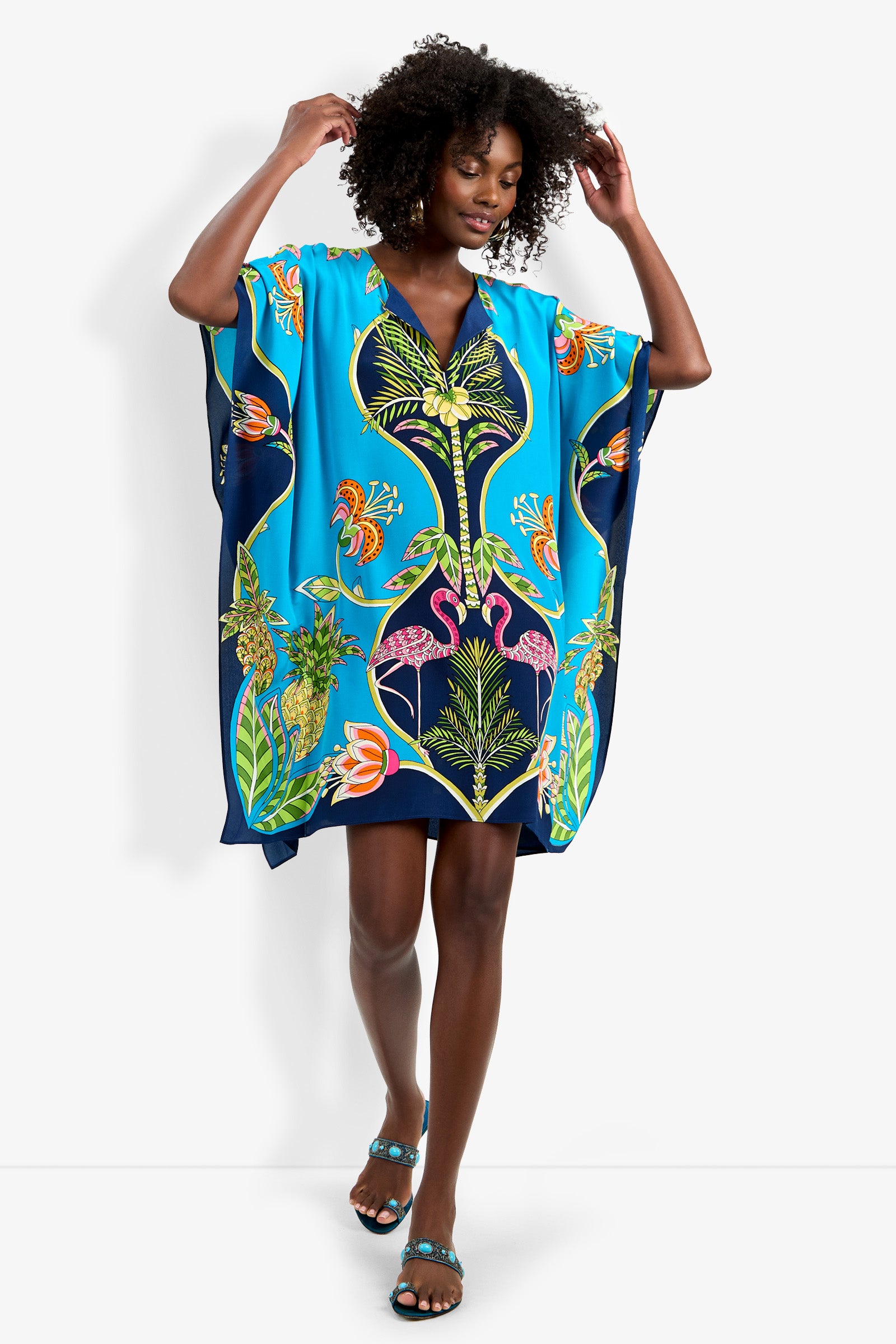 A woman, barefoot and smiling, poses with raised arms in the Theodora Caftan Dress—a loose, vibrant blue dress adorned with colorful tropical prints of flamingos, leaves, and flowers—set against a plain background.