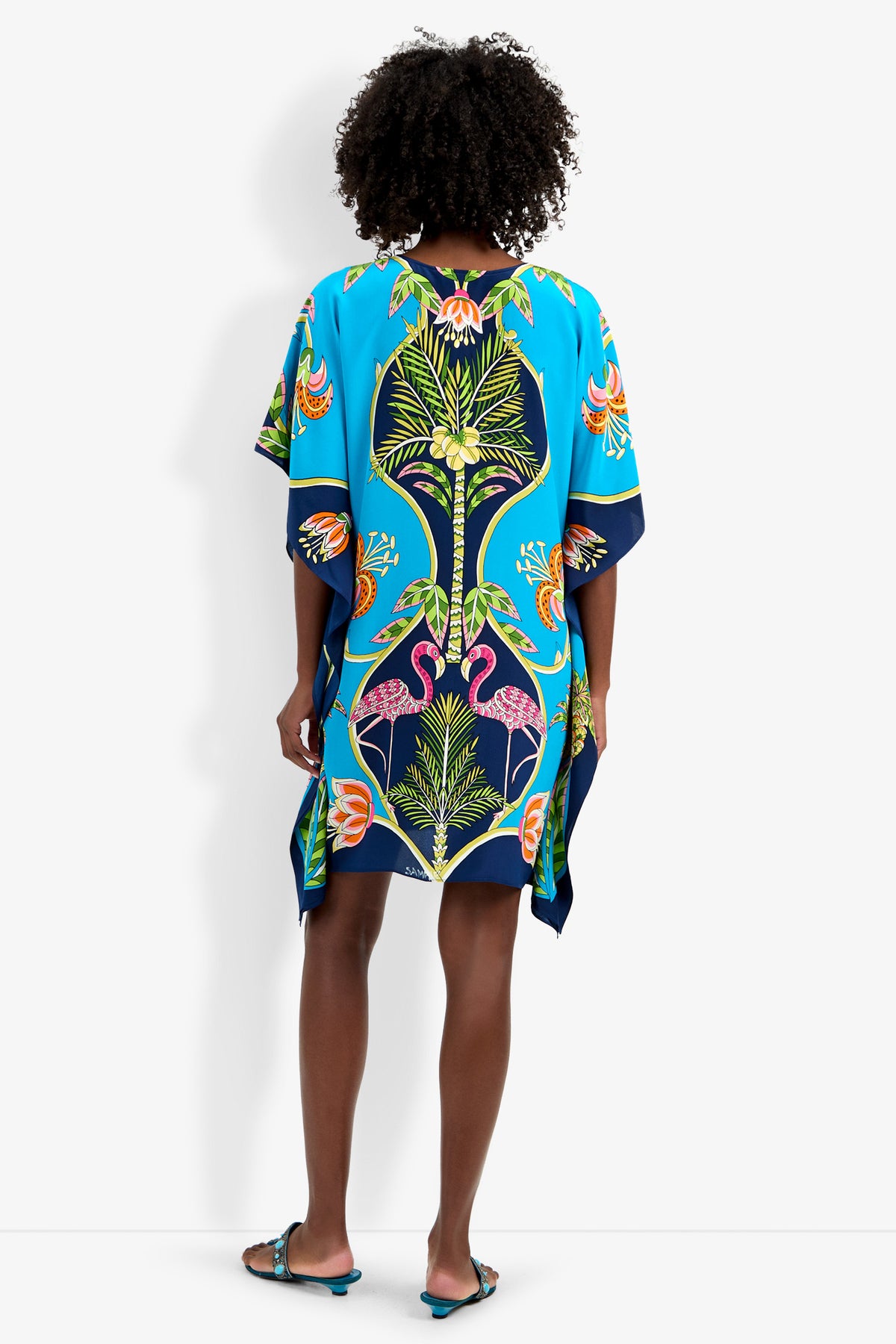 A person faces away, wearing Theodora Caftan Dress—a loose, vibrant blue piece with colorful botanical and floral patterns. The dress features wide sleeves, falls just above the knees, and is paired with flat sandals.