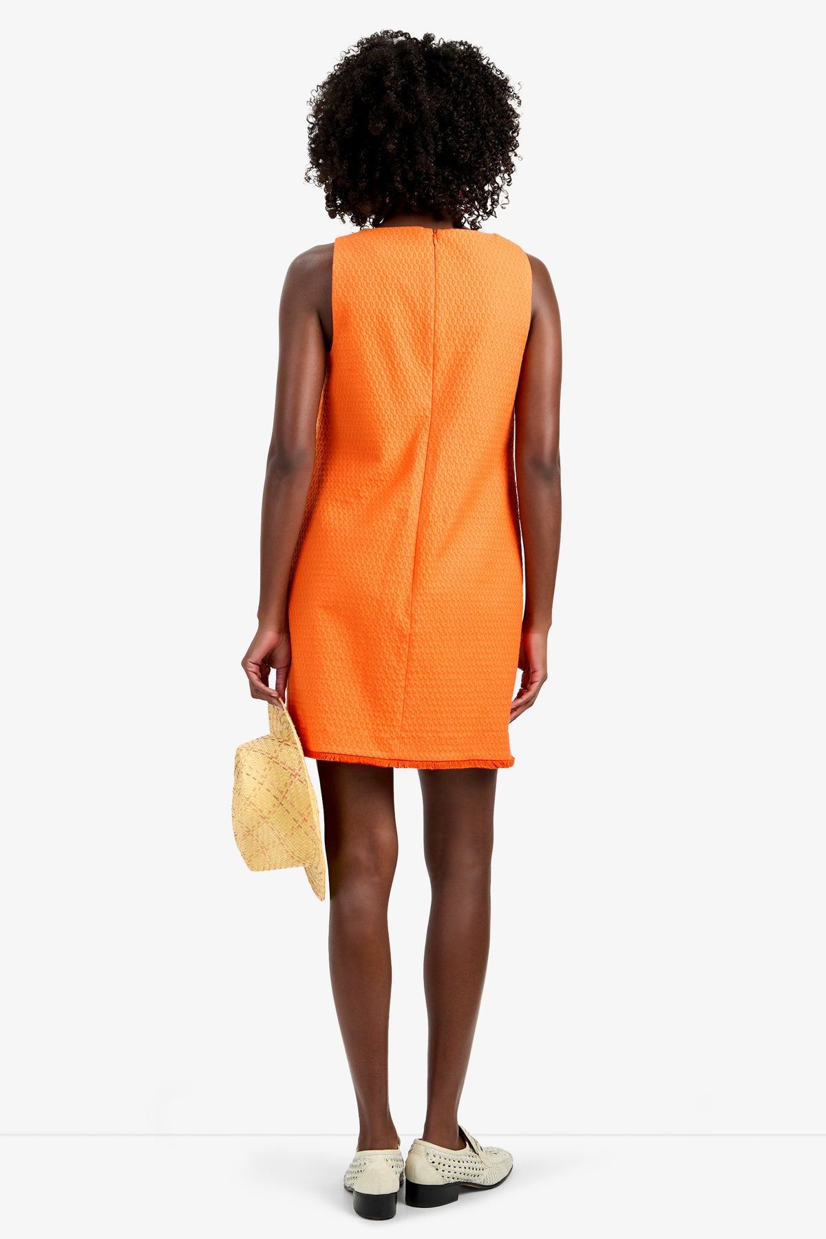 A person with curly hair, seen from behind, wears the WAVESON DRESS—an orange sleeveless viscose dress—with white shoes and holds a straw hat in their left hand against a plain white background.