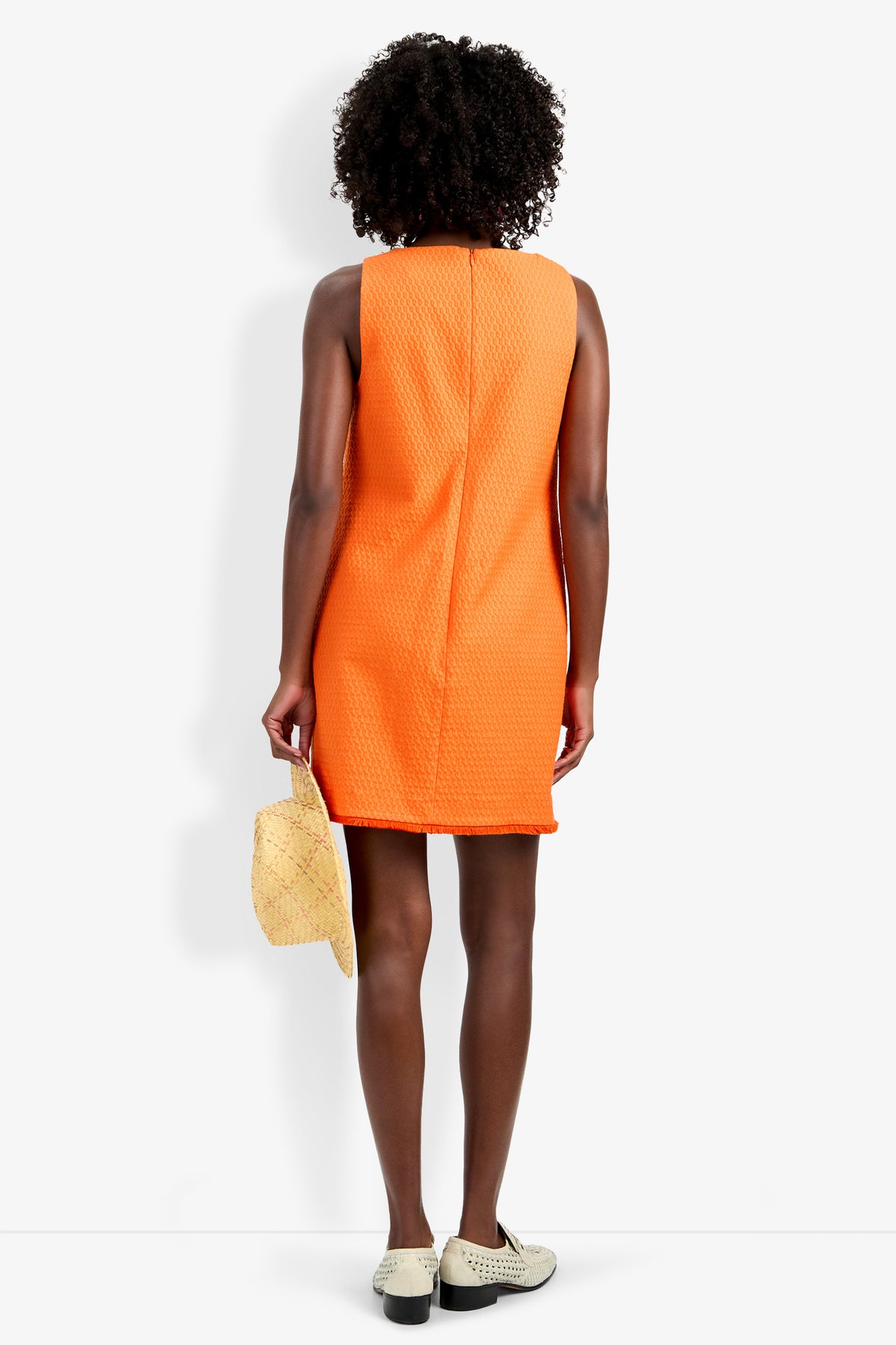 A person with curly hair is seen from the back, wearing the sleeveless orange Waveson Dress and white shoes, holding a straw hat in their left hand against a plain white background.