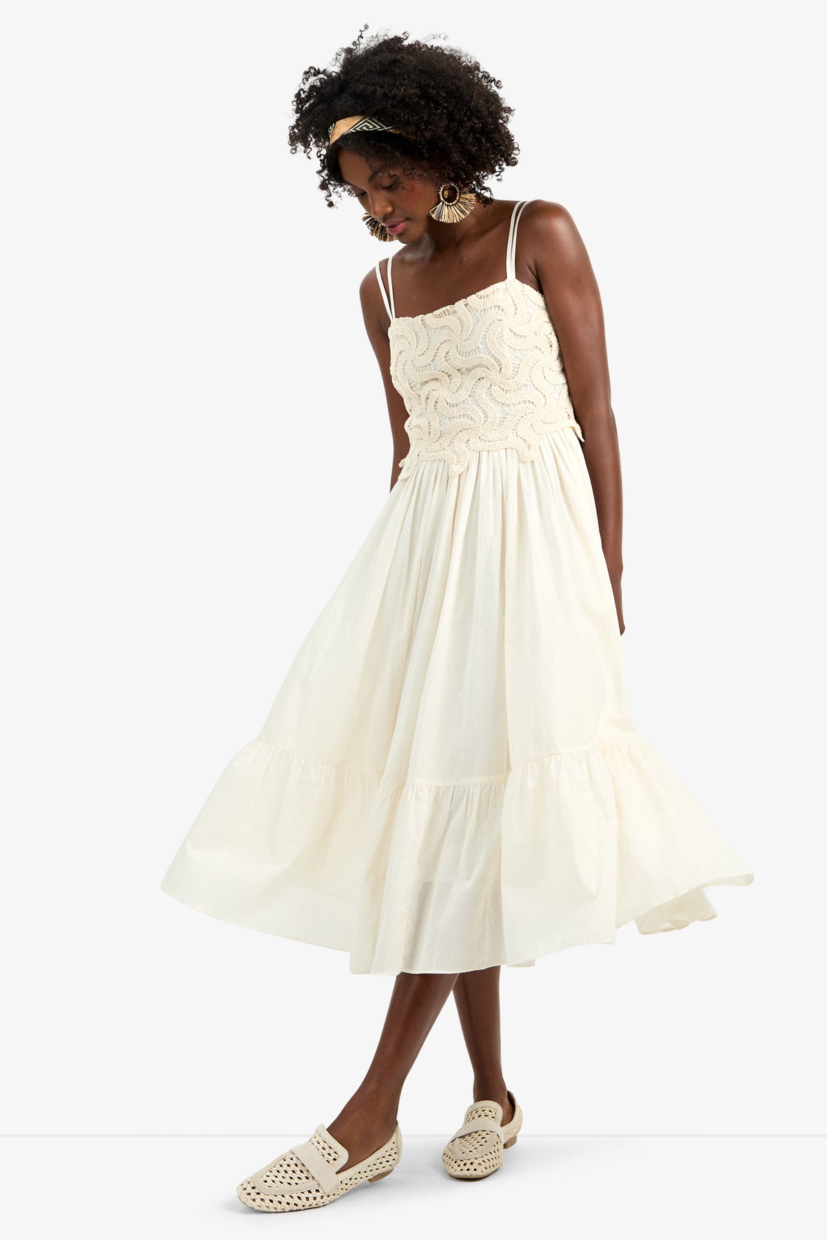 A woman stands against a plain background wearing the BAYO DRESS, a cream sleeveless 100% cotton dress with a textured top and ruffled skirt, paired with cream woven slip-on shoes, large earrings, and a yellow headband.
