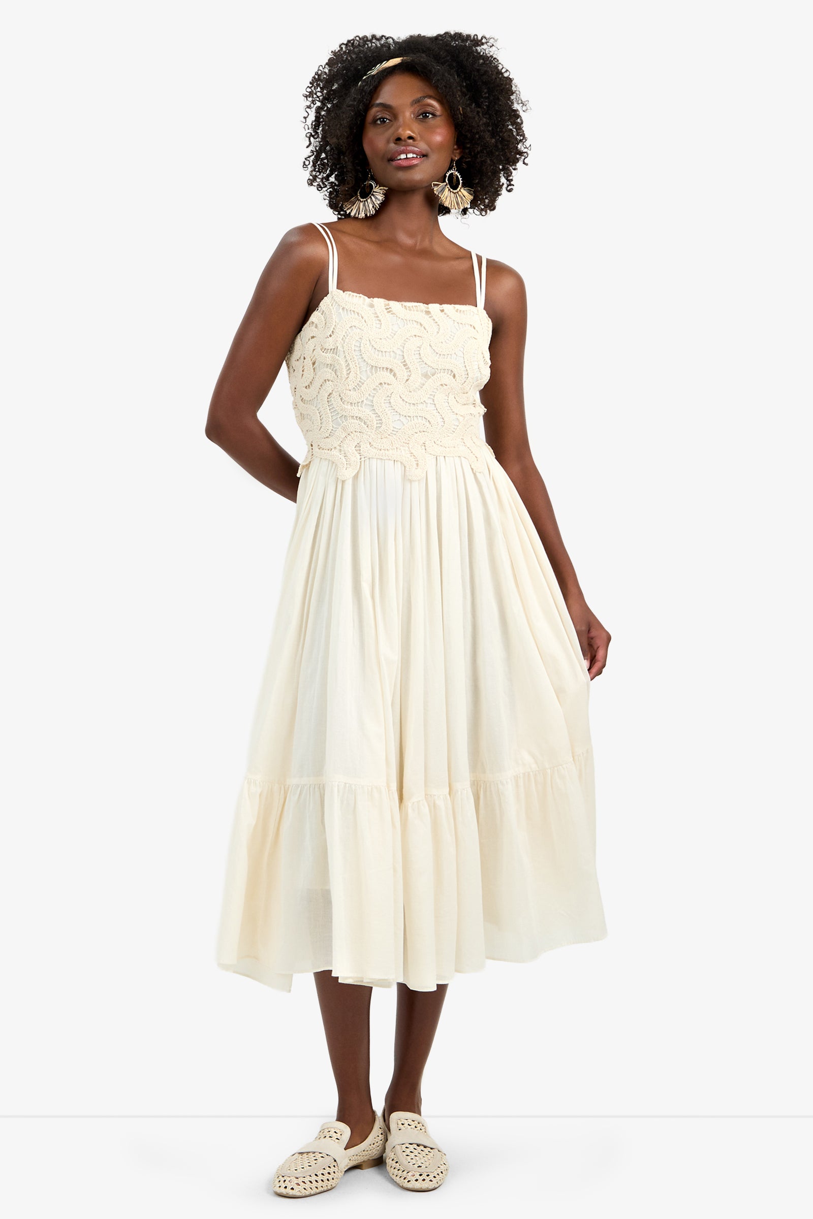A woman in the BAYO DRESS—a cream, knee-length 100% cotton dress with a textured bodice, spaghetti straps, and a flowy skirt—wears large earrings, woven shoes, and has curly hair.