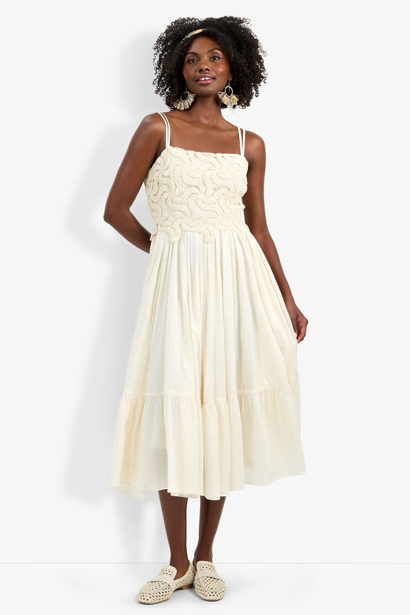 A woman in the Bayo Dress—sleeveless, cream, with a textured wavy bodice and flowing midi skirt—paired with statement earrings, curly hair, and cream woven slip-on shoes.