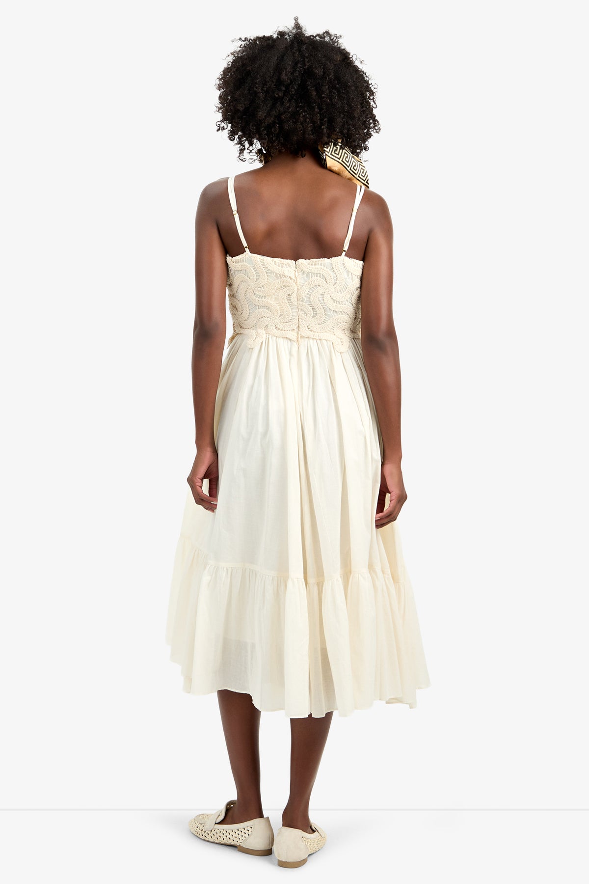 A woman with curly hair faces away, wearing the BAYO DRESS—a 100% cotton, cream-colored, sleeveless knee-length dress with a fitted bodice and flowing skirt—paired with flat light shoes and a patterned scarf around her neck.