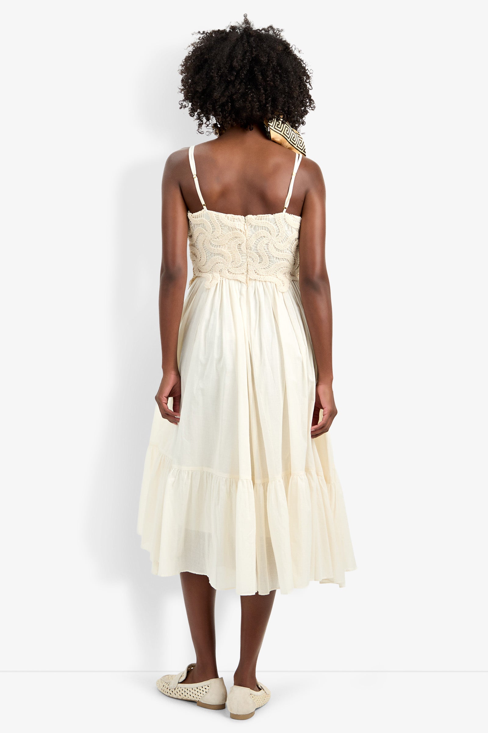 A person with curly hair wears the Bayo Dress, a cream sleeveless knee-length dress, paired with flat shoes, standing facing away from the camera against a plain white background.