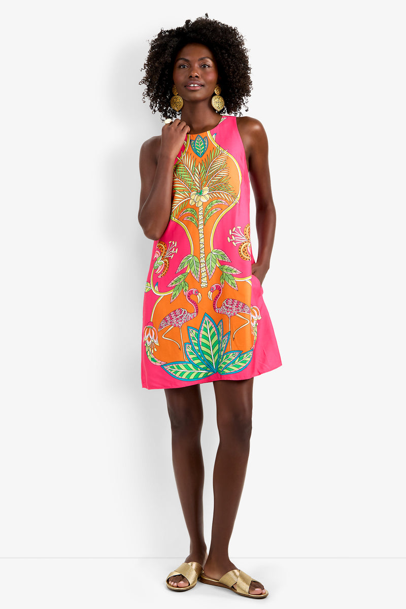 Woman standing and smiling against a white background, wearing the Loveage Dress—a sleeveless, bright pink dress with a tropical leaf and flower print—paired with gold earrings and gold sandals.